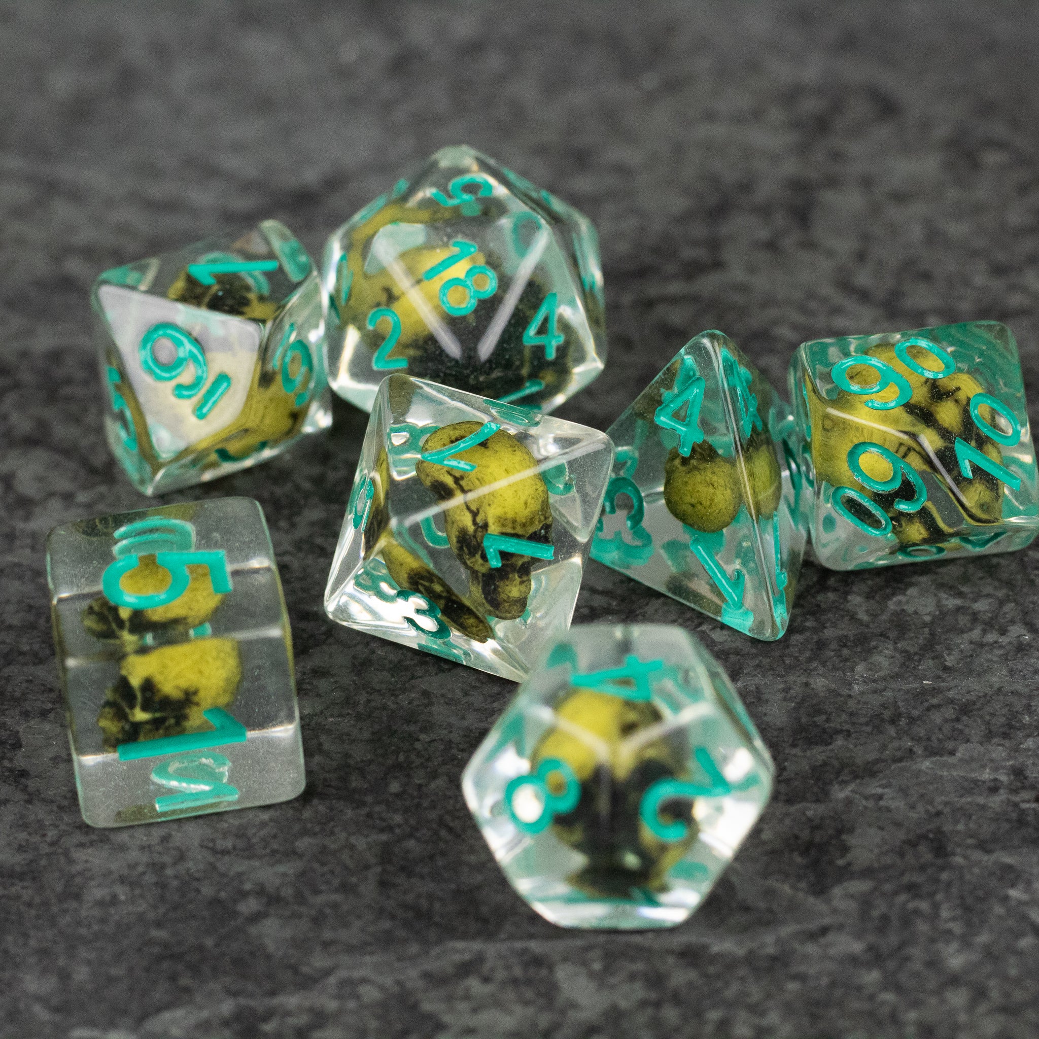 Yellow Skull Dice Set - Clear polyhedral dice set with embedded yellow skulls and teal numbers on gray surface.