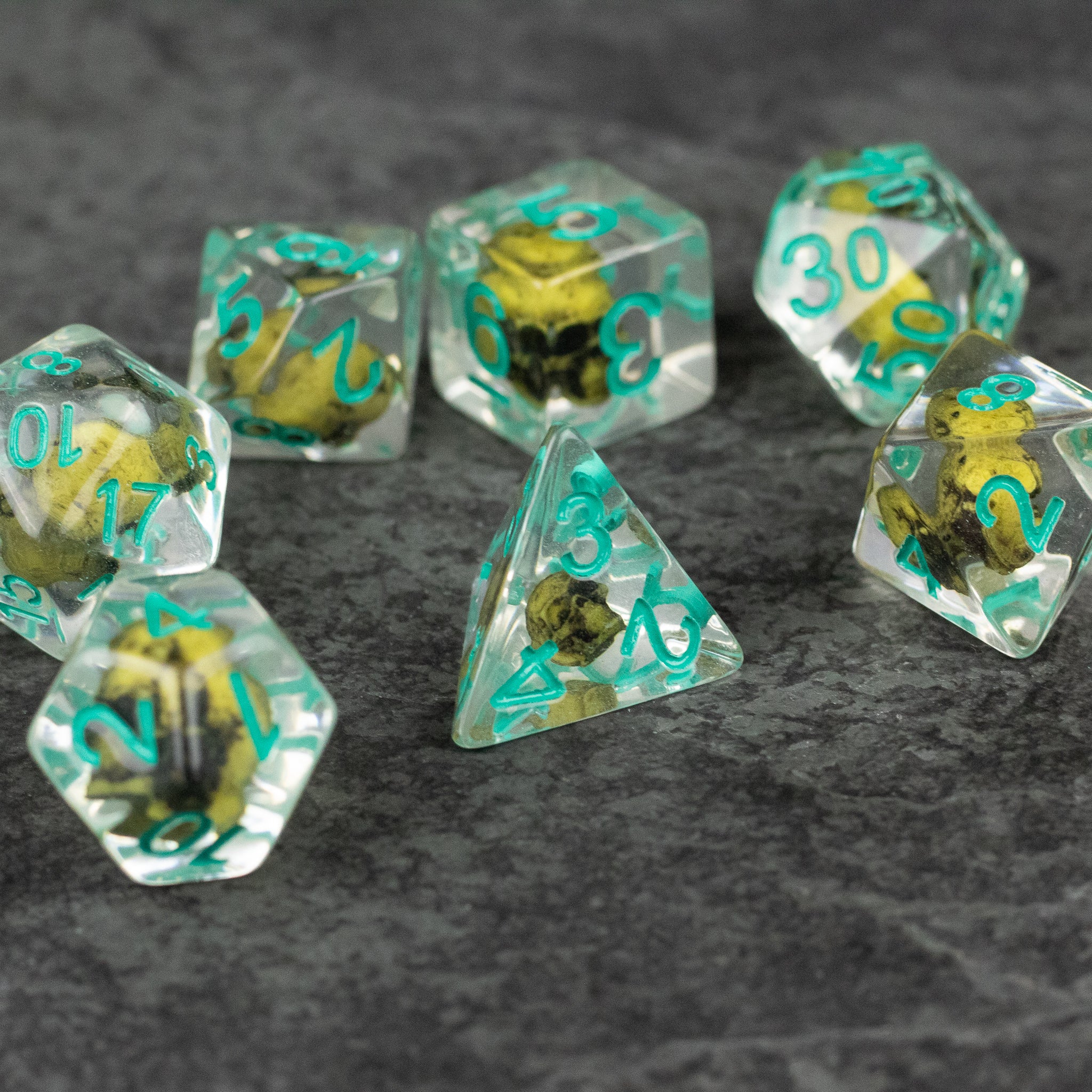 Yellow Skull Dice Set - Set of seven transparent polyhedral dice with yellow skulls inside and teal numbers.
