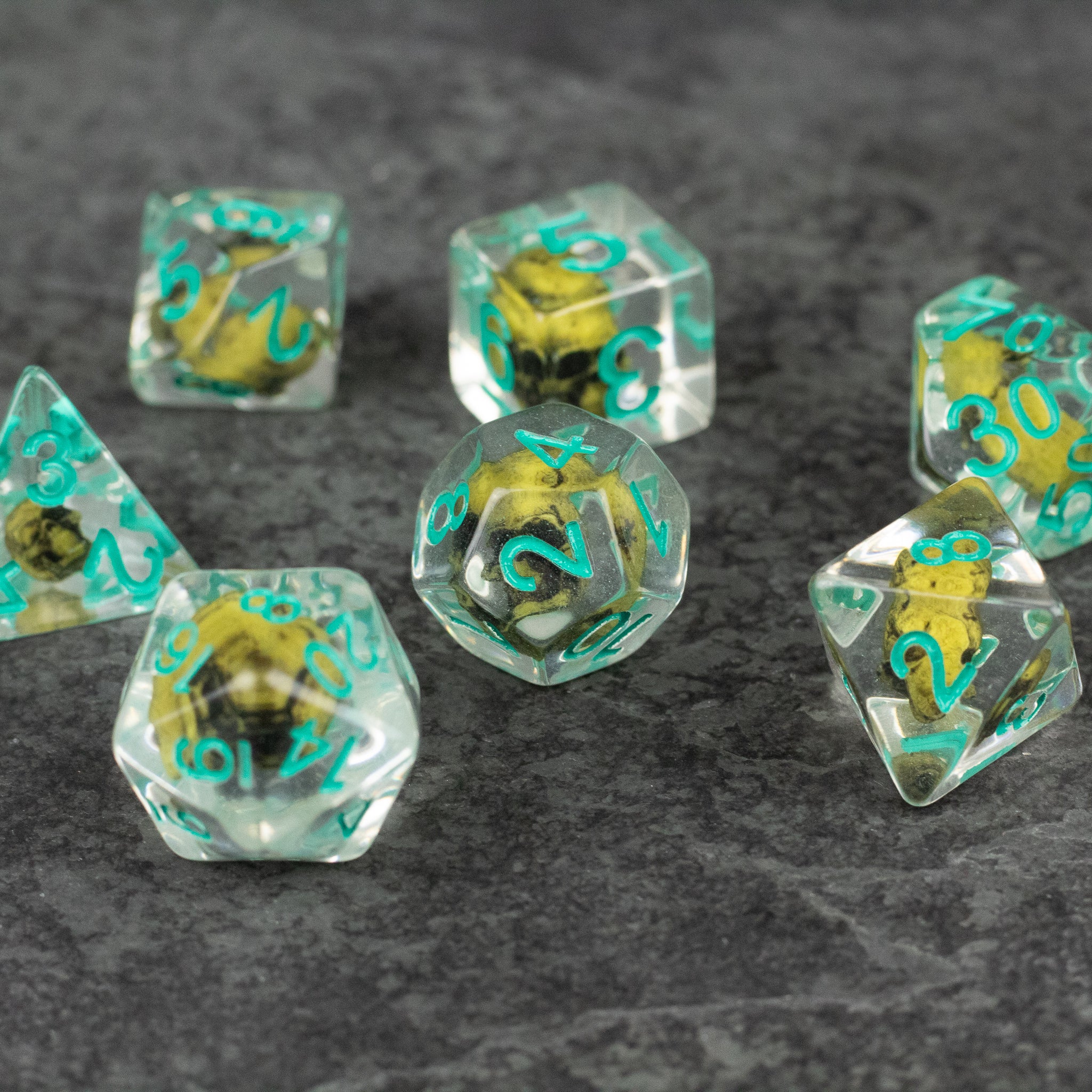 Yellow Skull Dice Set - Set of seven transparent dice with yellow skulls inside and teal numbers on a gray surface.