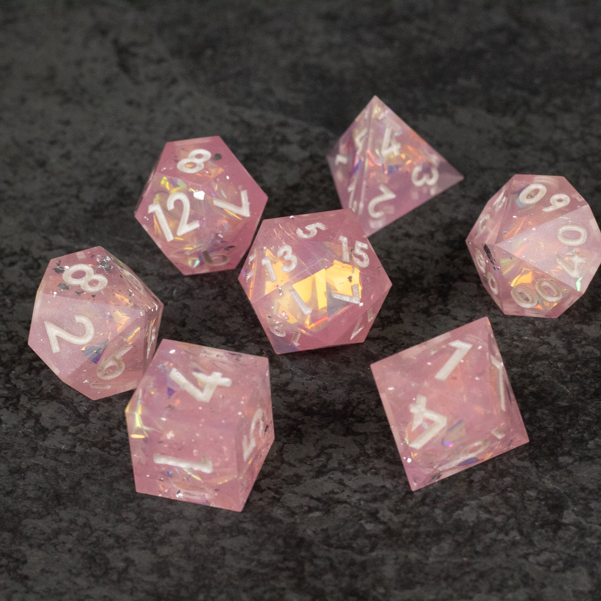 Bubblegum Dice Set - Seven pink translucent sharp-edged polyhedral dice with white numbers and a gold die in the center on a dark surface.