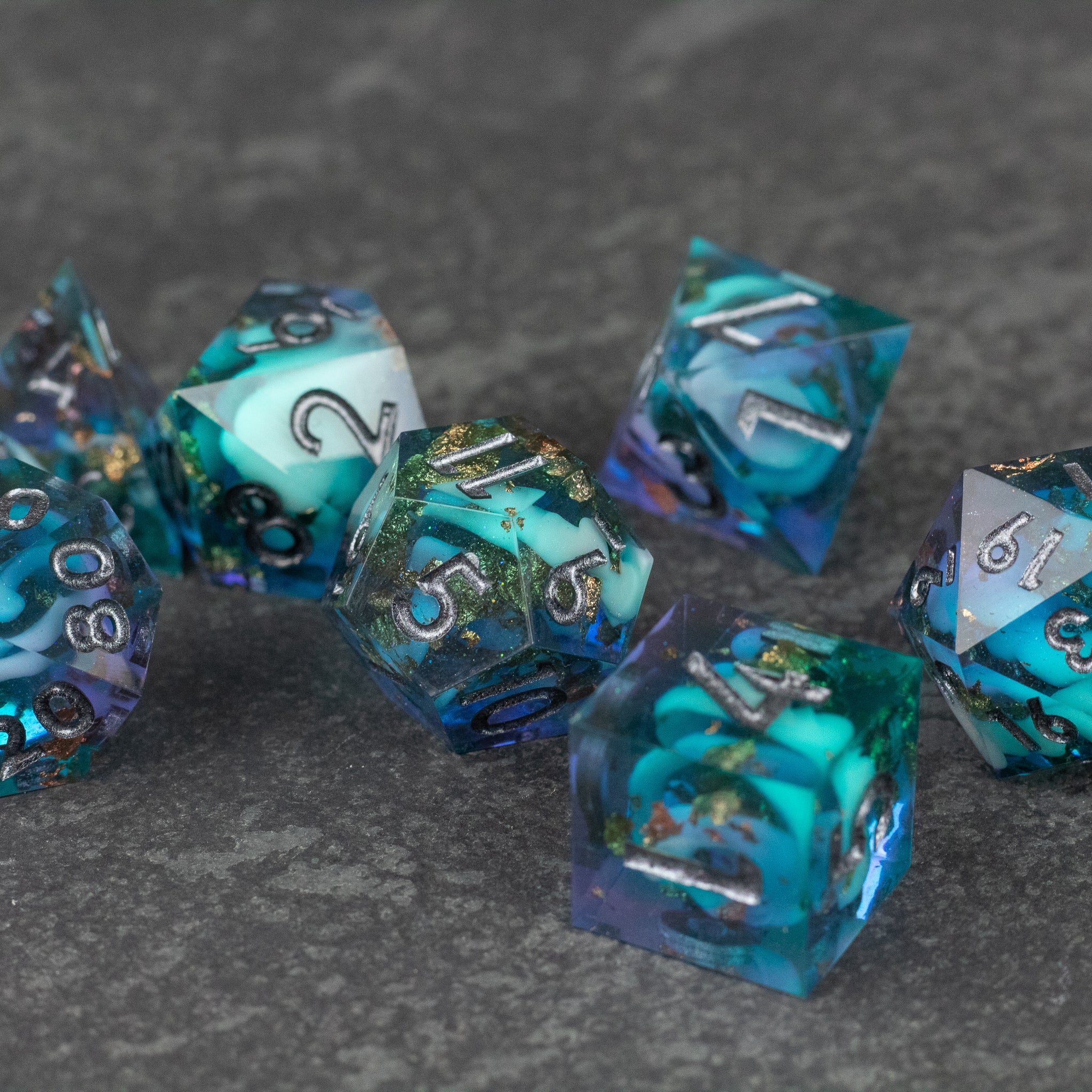 Frostfall Dice Set - Set of translucent blue and teal polyhedral dice with sharp edges and metallic silver numbers on a gray textured surface.