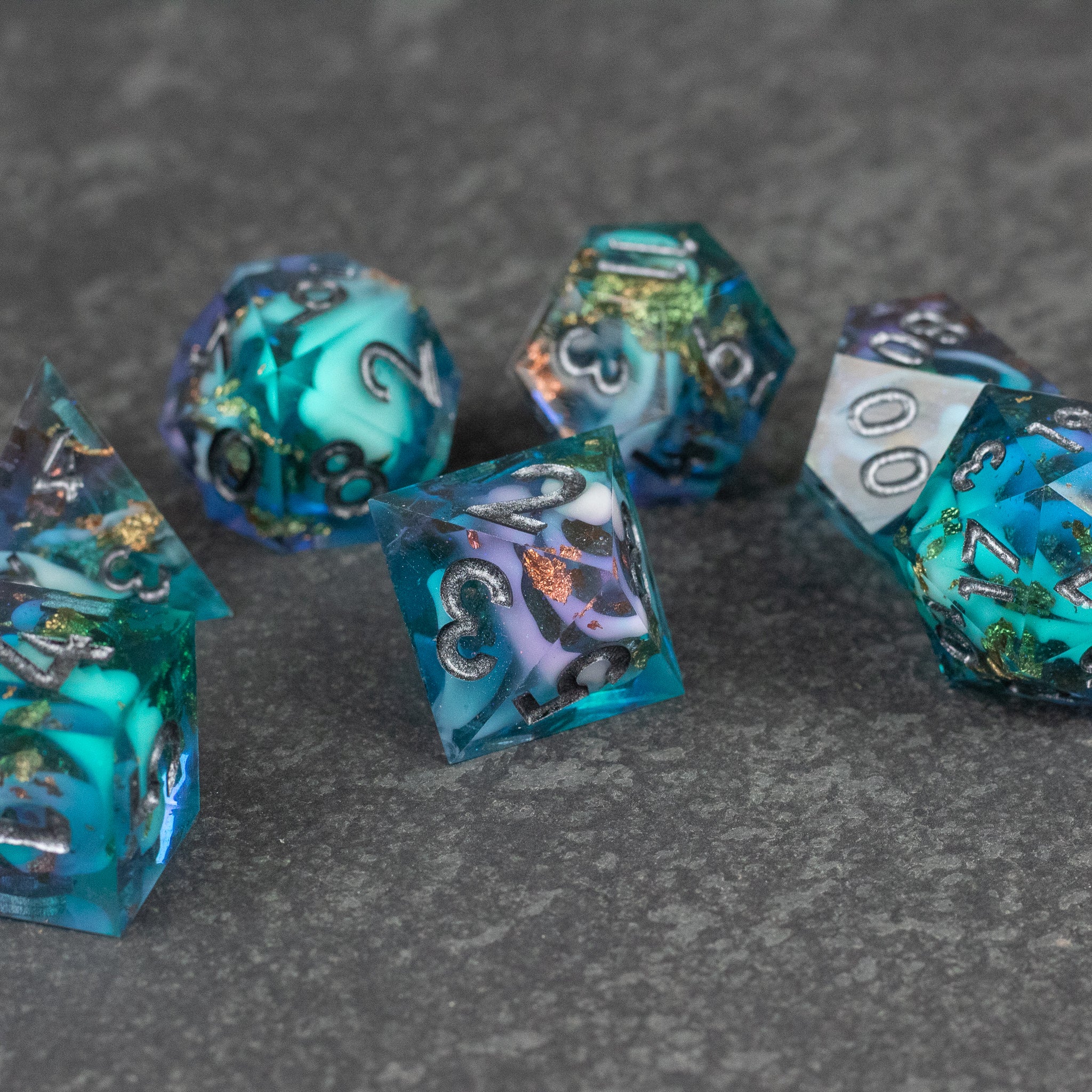 Frostfall Dice Set - Translucent blue and teal sharp-edged dice set with silver numbering and swirling icy inclusions on gray stone surface.