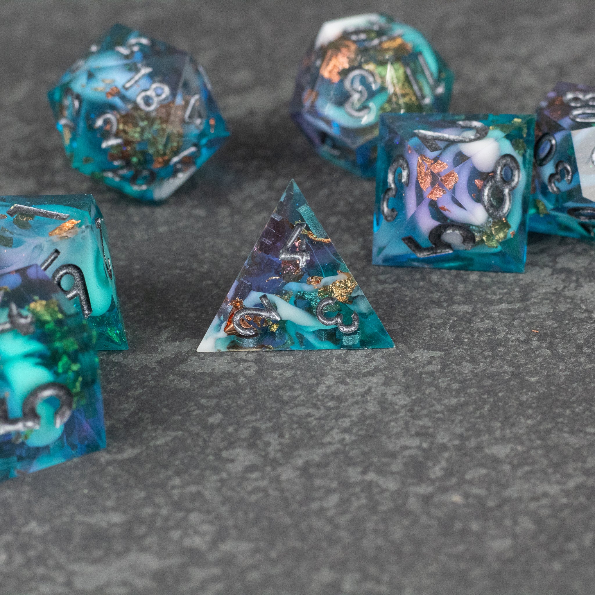 Frostfall Dice Set - Blue and green sharp edge polyhedral dice set with swirling orange and white details on gray stone background.