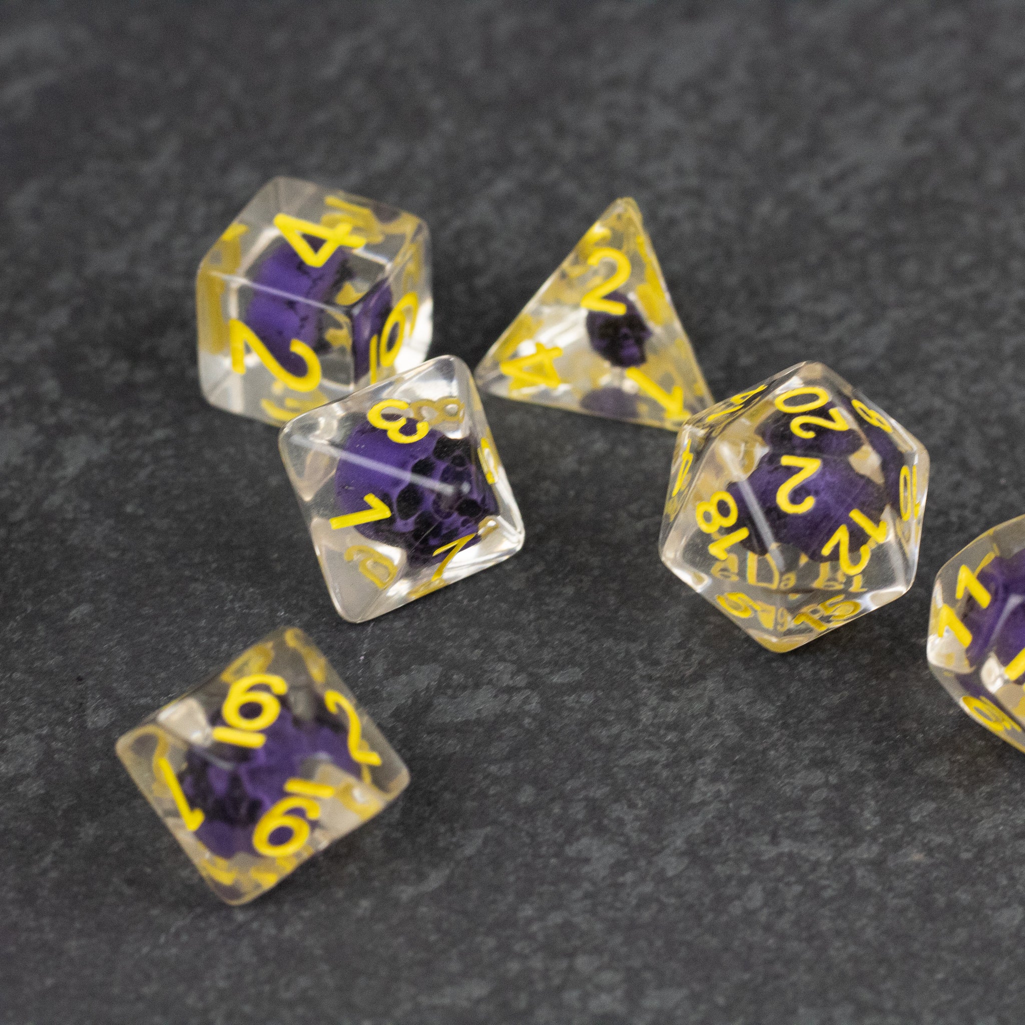 Purple Skull Dice Set - Set of six transparent dice with purple skulls inside and yellow numbering on black surface.