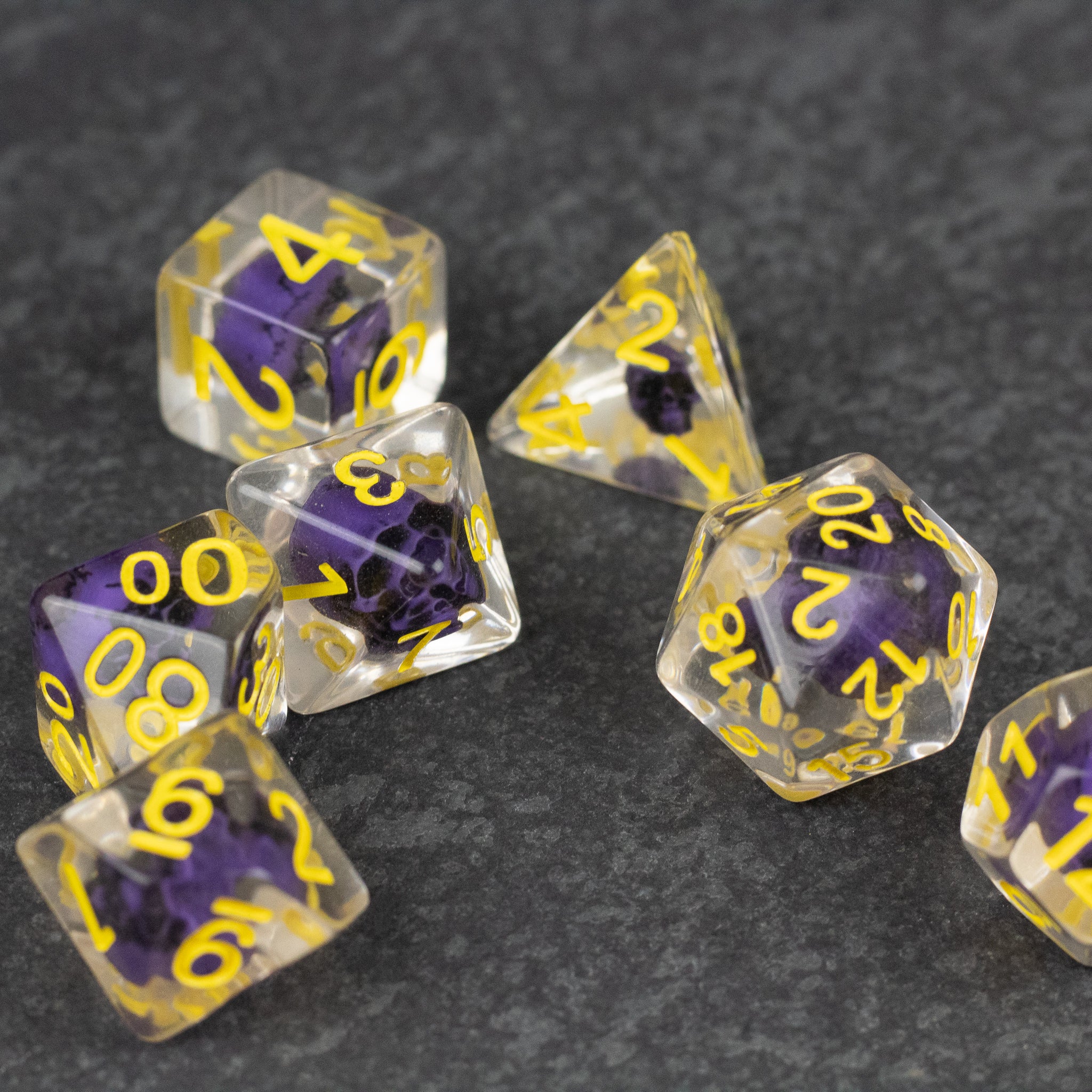Purple Skull Dice Set - Set of seven translucent dice with purple skulls inside and yellow numbers, including d20, d12, d10, d8, d6, and d4 shapes.