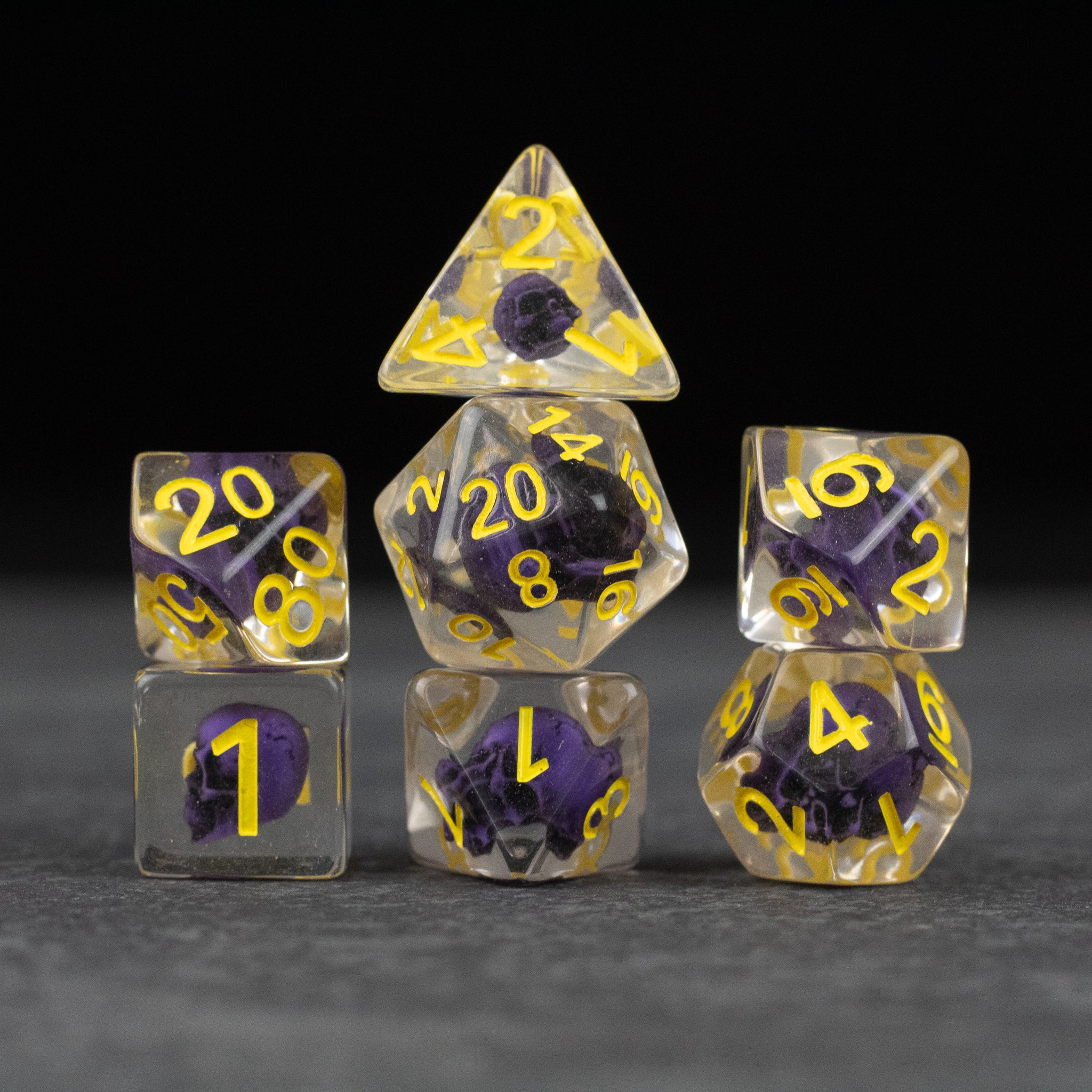 Purple Skull Dice Set - Seven transparent dice with embedded purple skulls and yellow numbering arranged in a pyramid shape on black background.