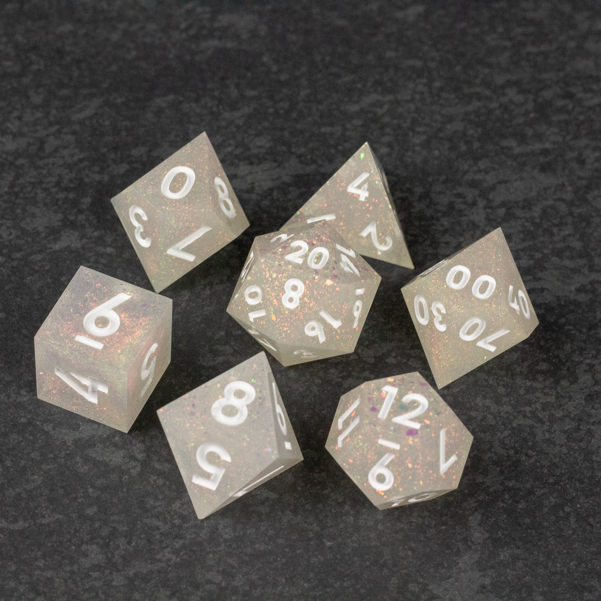 Snowfield Dice Set - Seven white sharp-edged polyhedral dice with clear speckled design and white numbers on a dark gray surface.