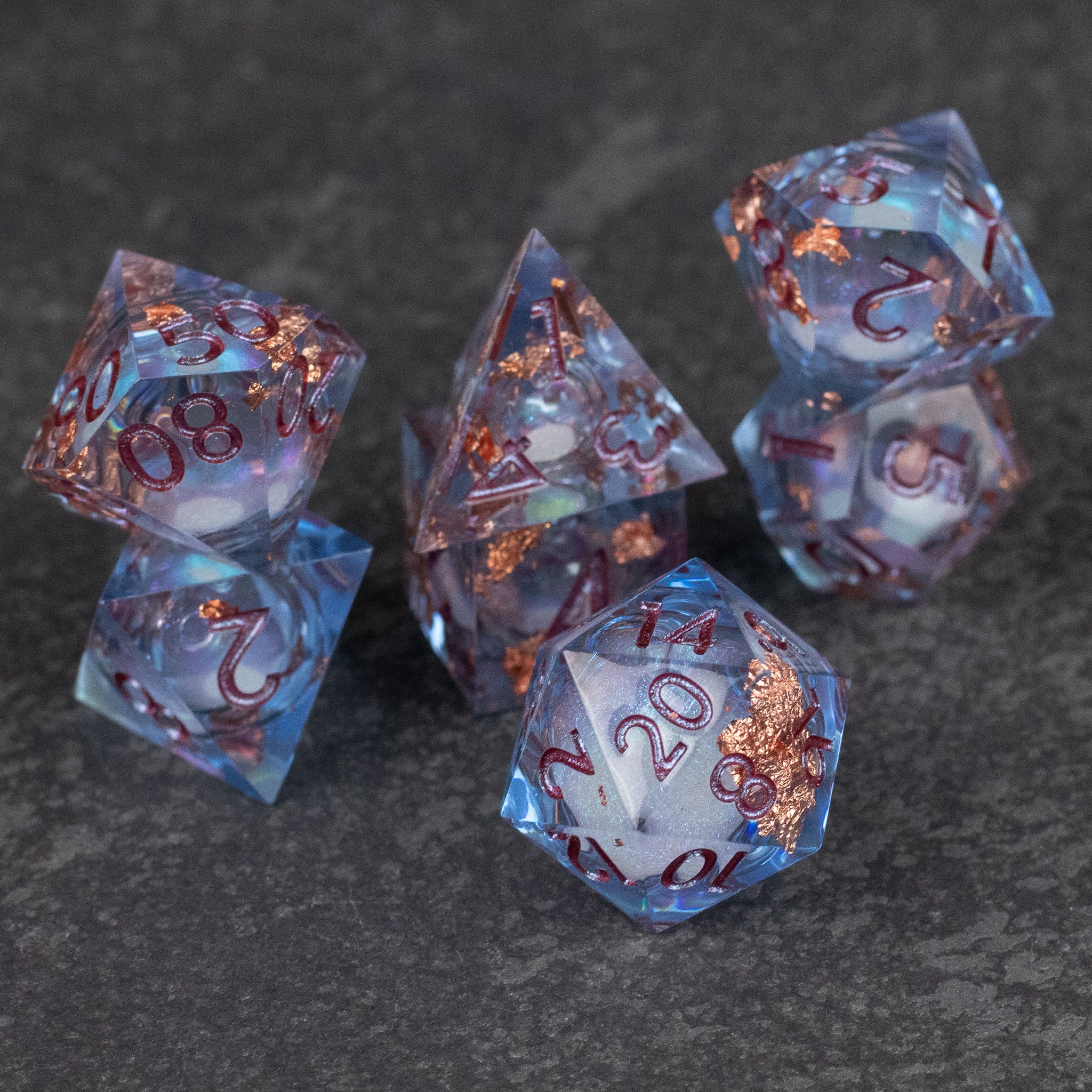 Temporal Rift Liquid Core Dice Set - Set of seven translucent blue sharp-edged dice with copper-colored liquid core and red numbers on dark stone surface.