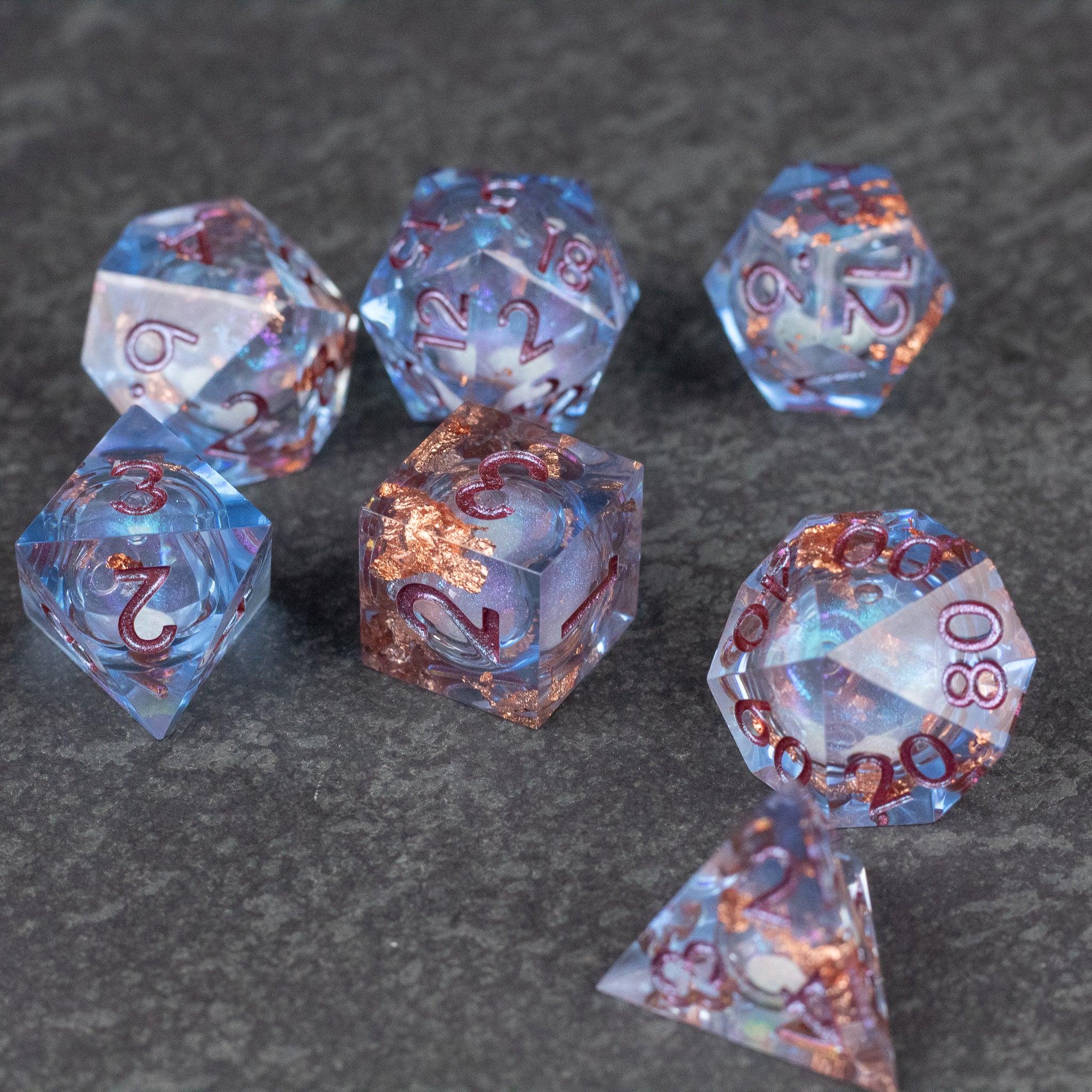 Temporal Rift Liquid Core Dice Set - Set of seven translucent blue liquid core polyhedral dice with sharp edges and copper-colored numbers on a dark textured.