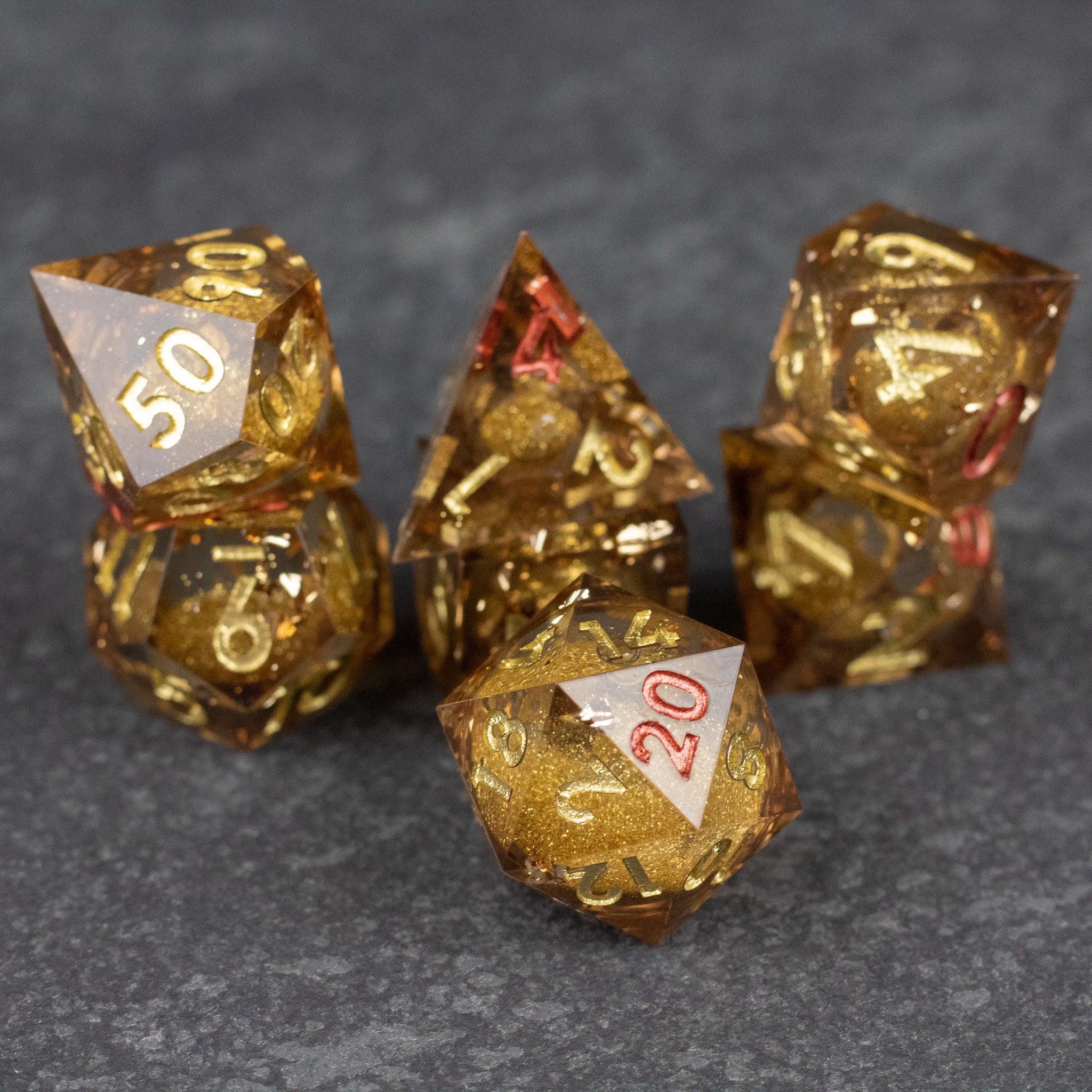 Dragon's Hoard Liquid Core Dice Set - Sharp-edged gold liquid core polyhedral dice set with white and red numbering on dark stone surface.