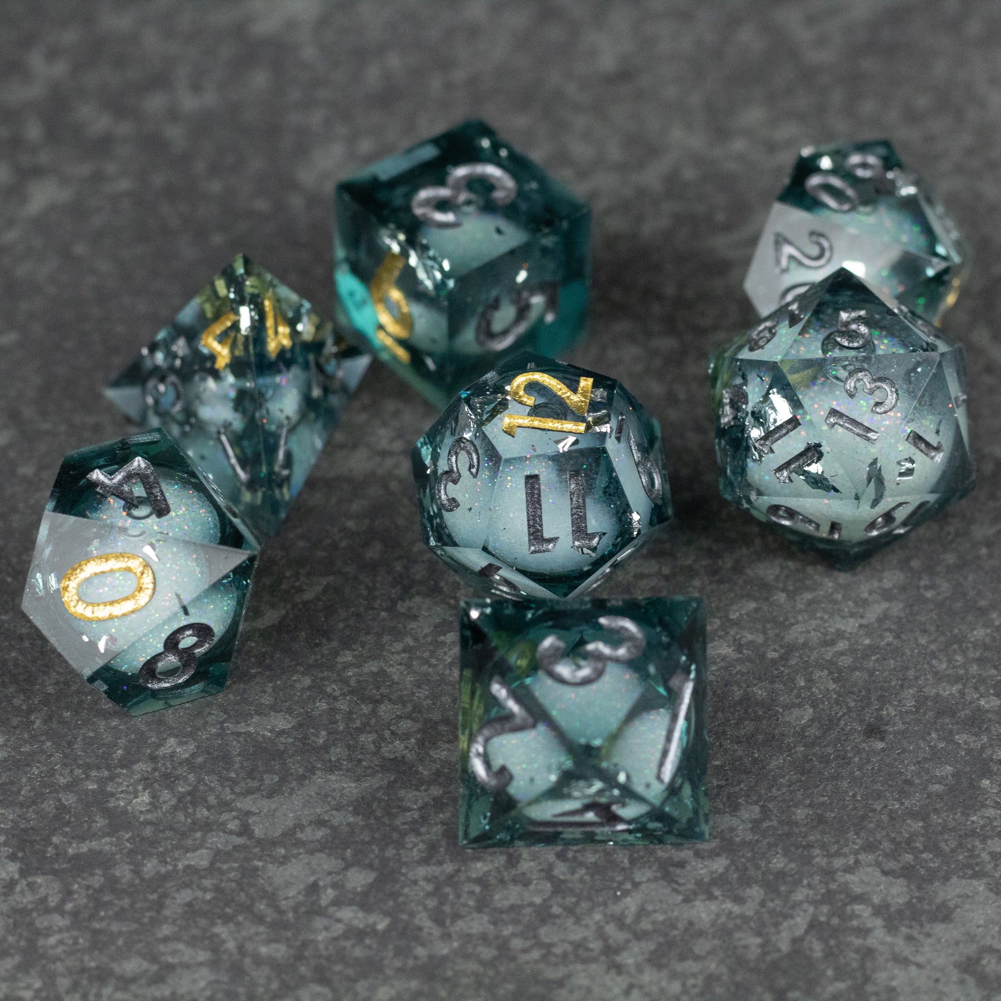 Call Of The Deep - Liquid Core Dice Set - Seven deep teal translucent dice with gold and white flecks, sharp edges, and white and gold numbers on gray textured.