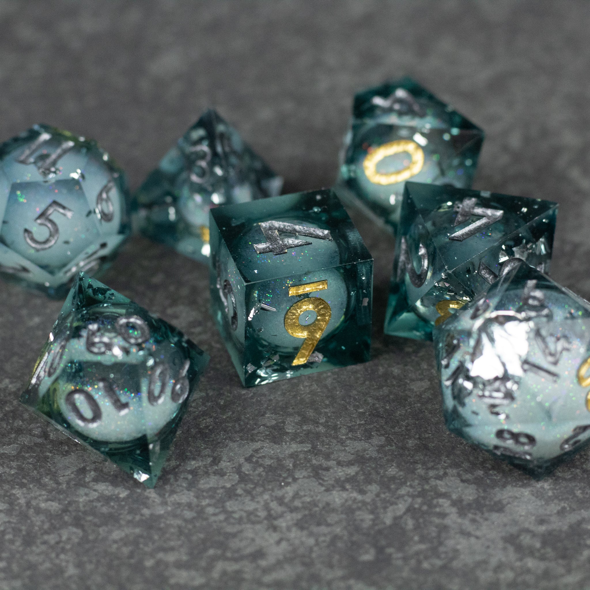 Call Of The Deep - Liquid Core Dice Set - Set of translucent dark teal sharp-edged dice with metallic silver liquid core and gold numbering.