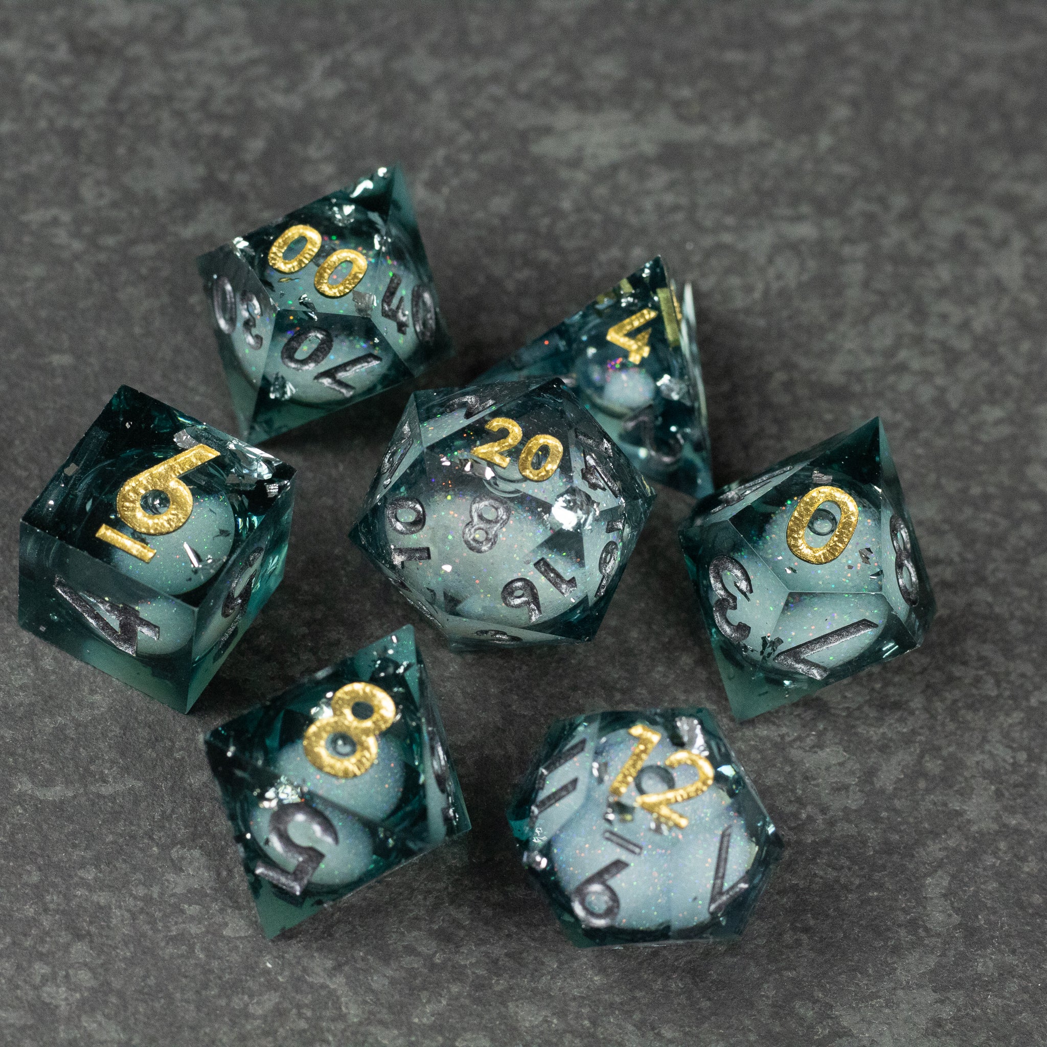 Call Of The Deep - Liquid Core Dice Set - Seven sharp-edged dice with transparent dark teal liquid cores and gold numbering on a gray surface.