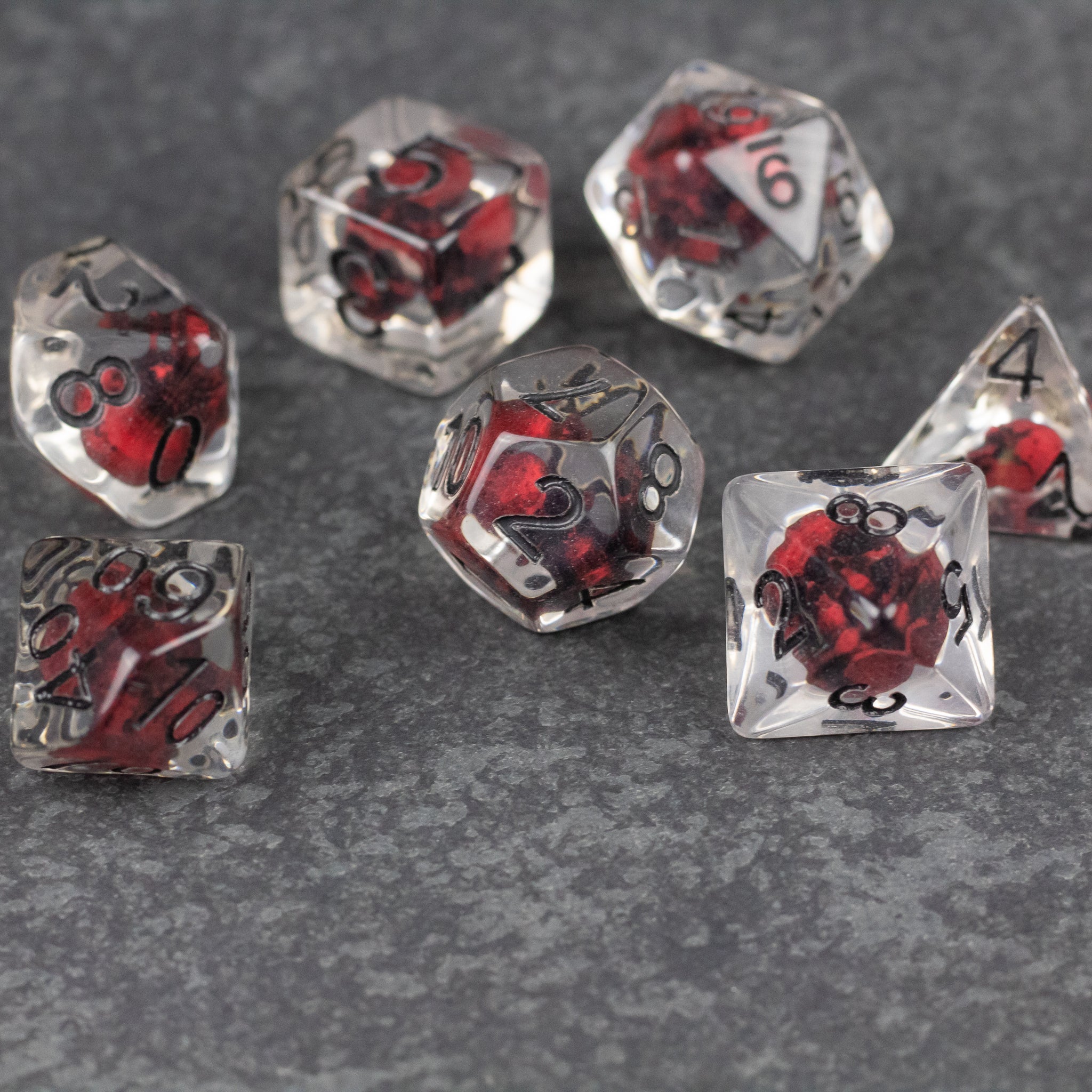 Red Skull Dice Set - Set of seven translucent dice with embedded red skulls and black numbering, various polyhedral shapes on dark textured.
