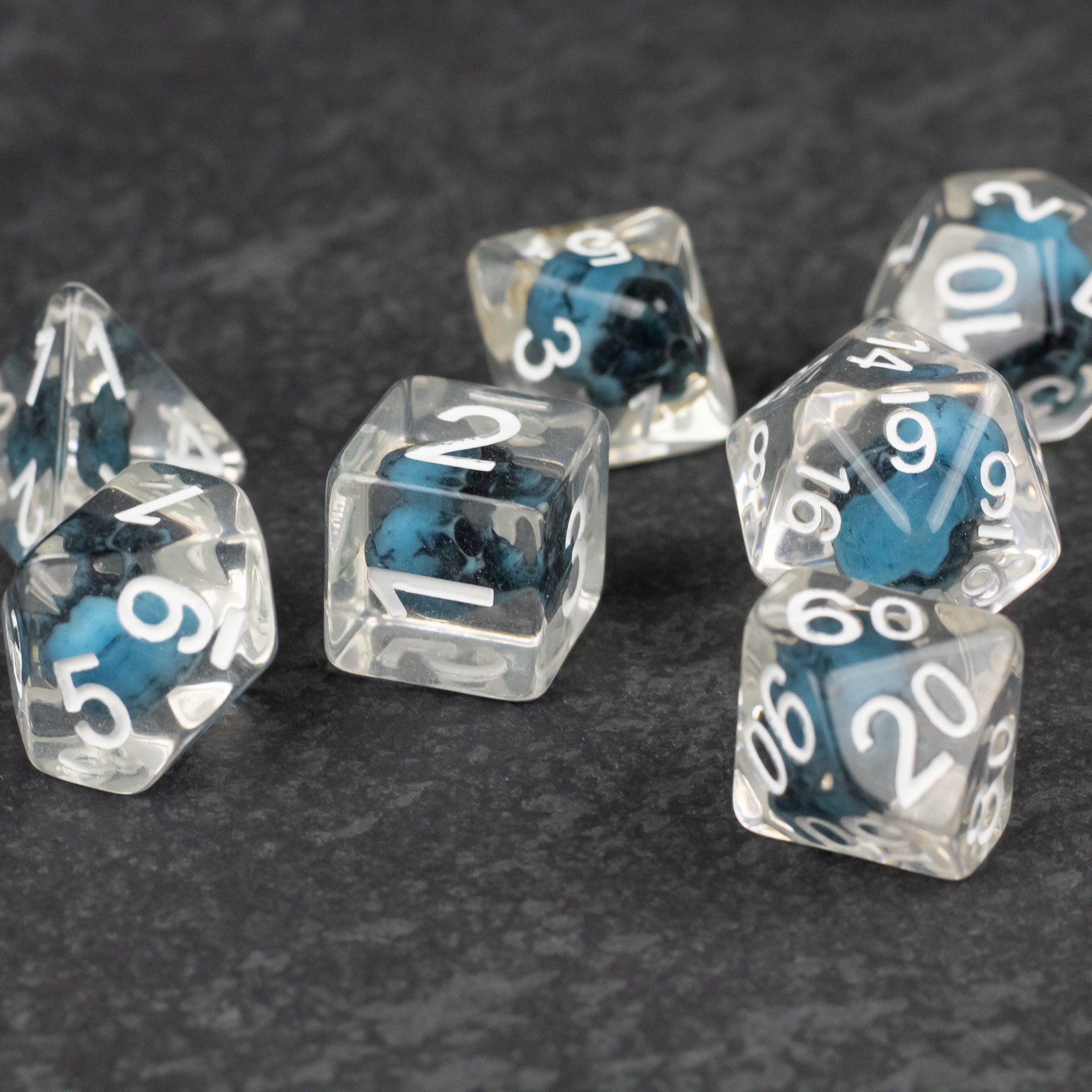 Blue Skull Dice Set - Set of clear resin dice with embedded blue skulls and white numbering on a dark textured surface.