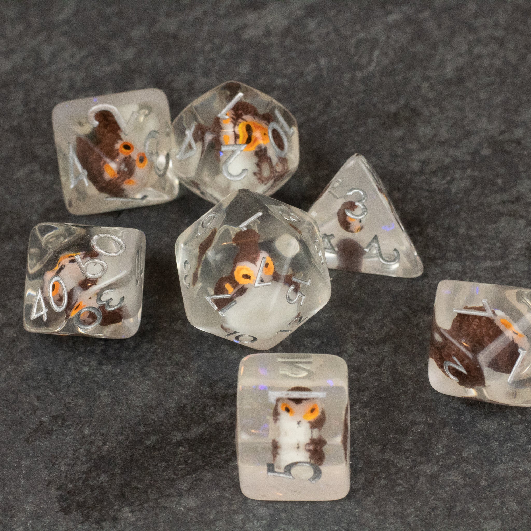 Owl Dice Set - Transparent polyhedral dice set with embedded miniature owls and orange numbering on a dark surface.