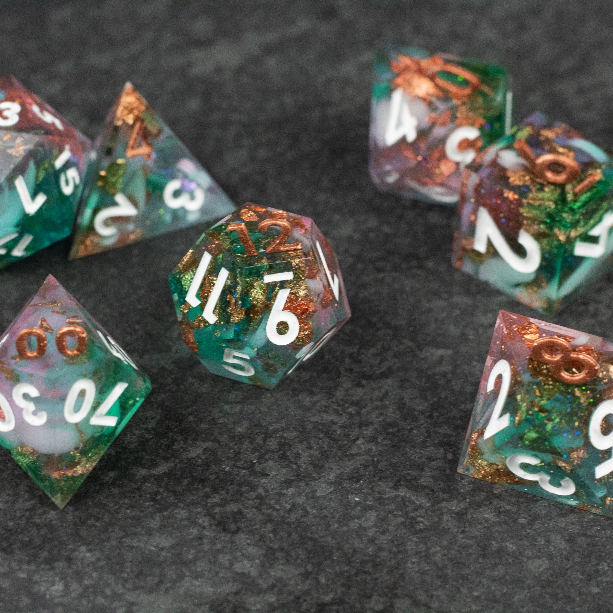 Scapeshift Dice Set - Seven translucent polyhedral dice in green and orange hues with white numbers and sharp edges on a dark surface.