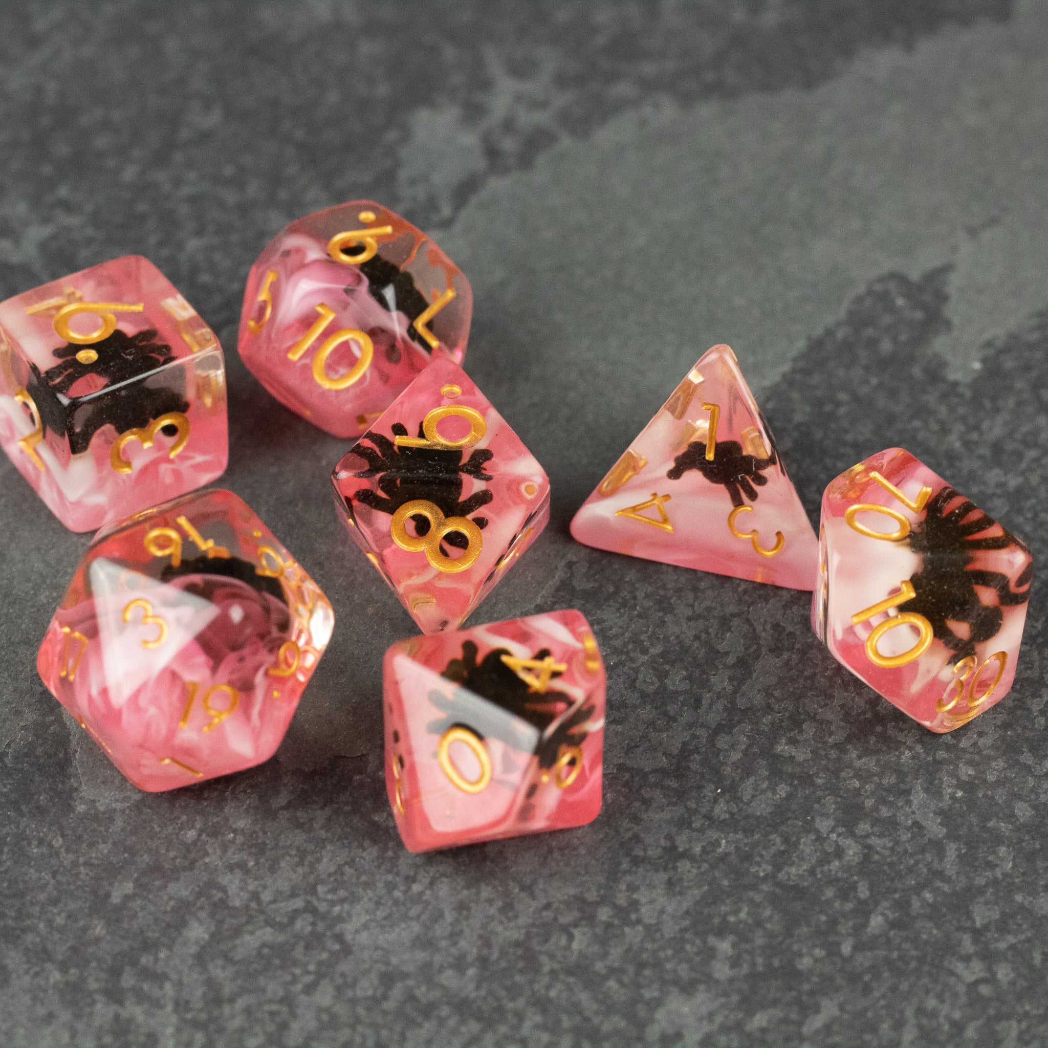 Black Spider Dice Set - Red - Seven red translucent polyhedral dice featuring black spider designs and gold numbers on a dark stone surface.