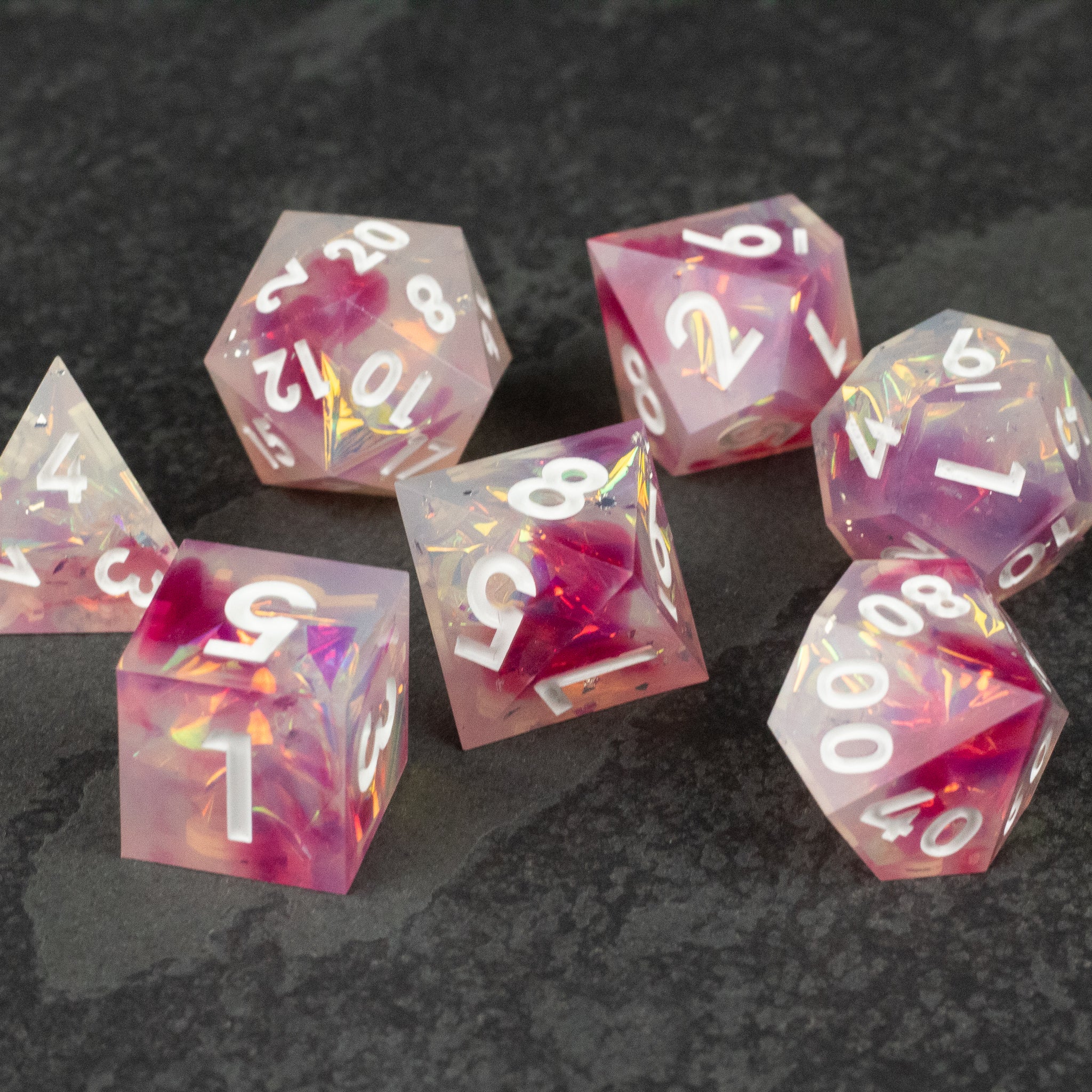 Lover's Kiss Dice Set - Set of seven translucent pink and white marbled polyhedral dice with white numbers and sharp edges on a dark surface.