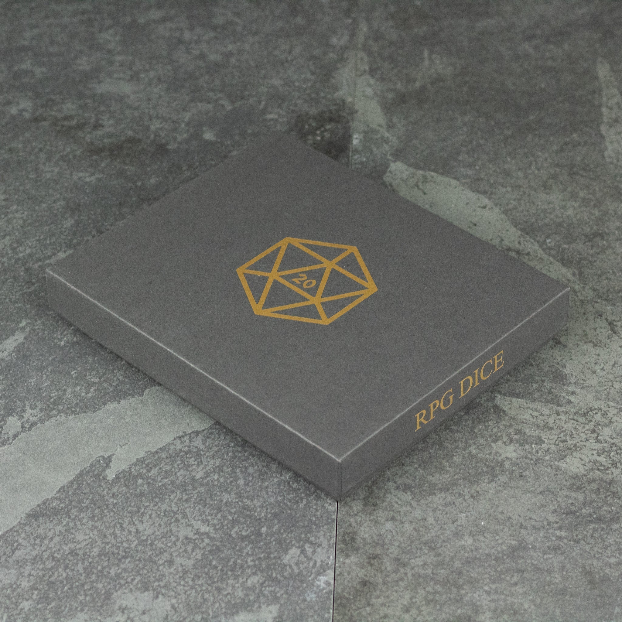 Metal Dice Set Gift Box - Closed gray gift box with gold geometric logo and "RPG Dice" text on side, resting on stone surface.