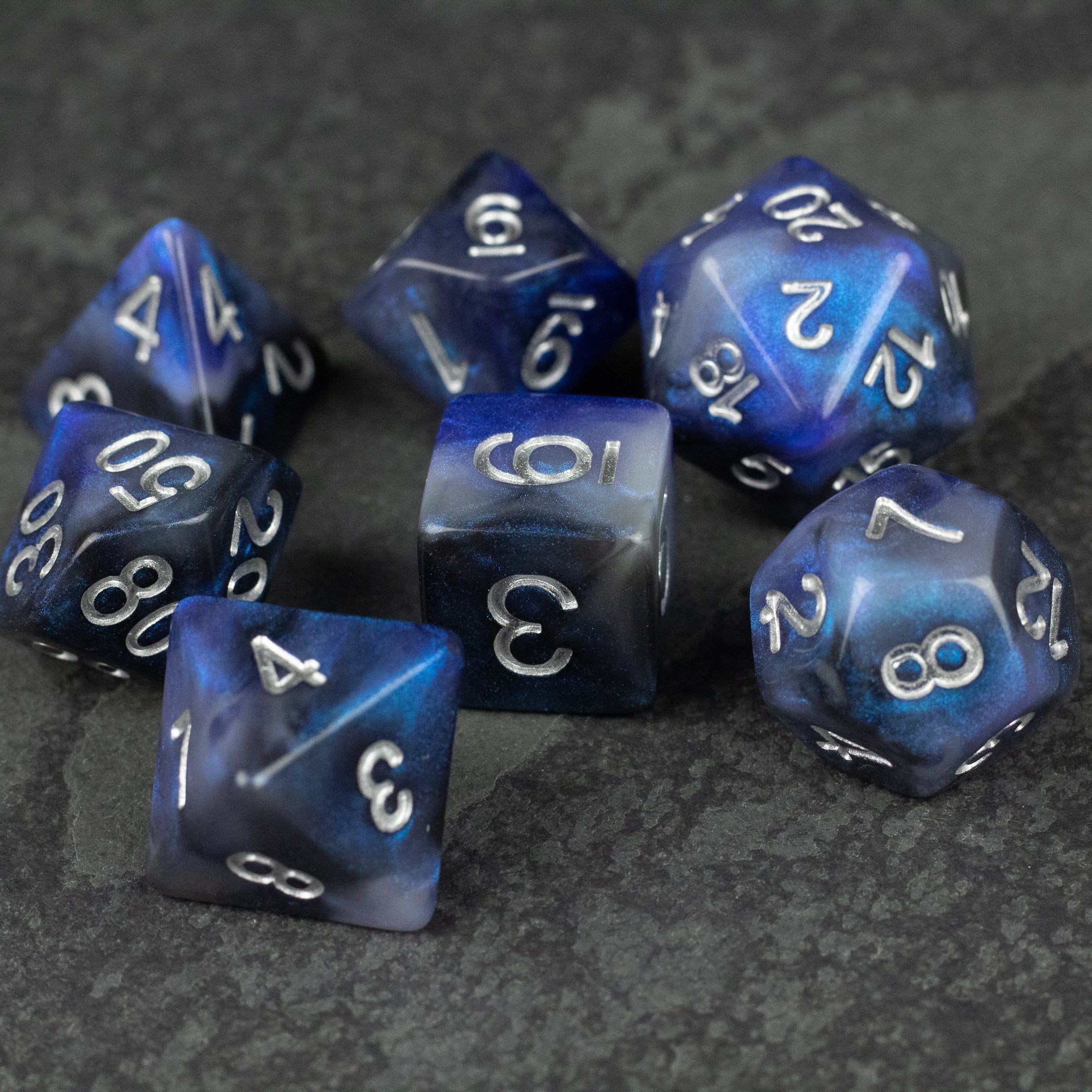 Cosmic Dust Dice Set - Set of seven blue and white marbled polyhedral dice with white numbers on a dark textured surface.