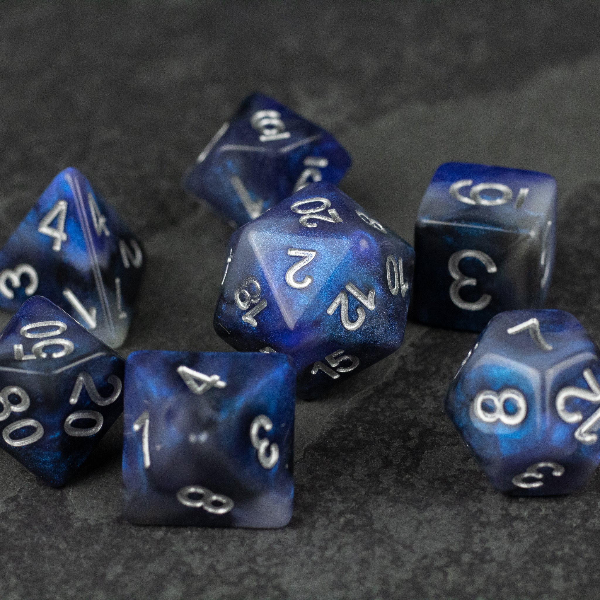 Cosmic Dust Dice Set - Set of seven blue dice with swirling cosmic patterns and white numbers, including d4, d6, d8, d10, d12, and d20 shapes.