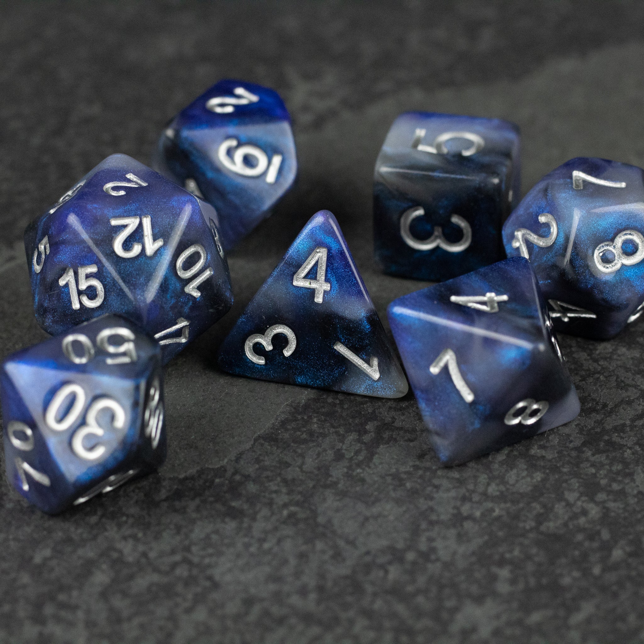 Cosmic Dust Dice Set - Set of seven glossy blue polyhedral dice with white numbering, featuring a marbled cosmic dust pattern.