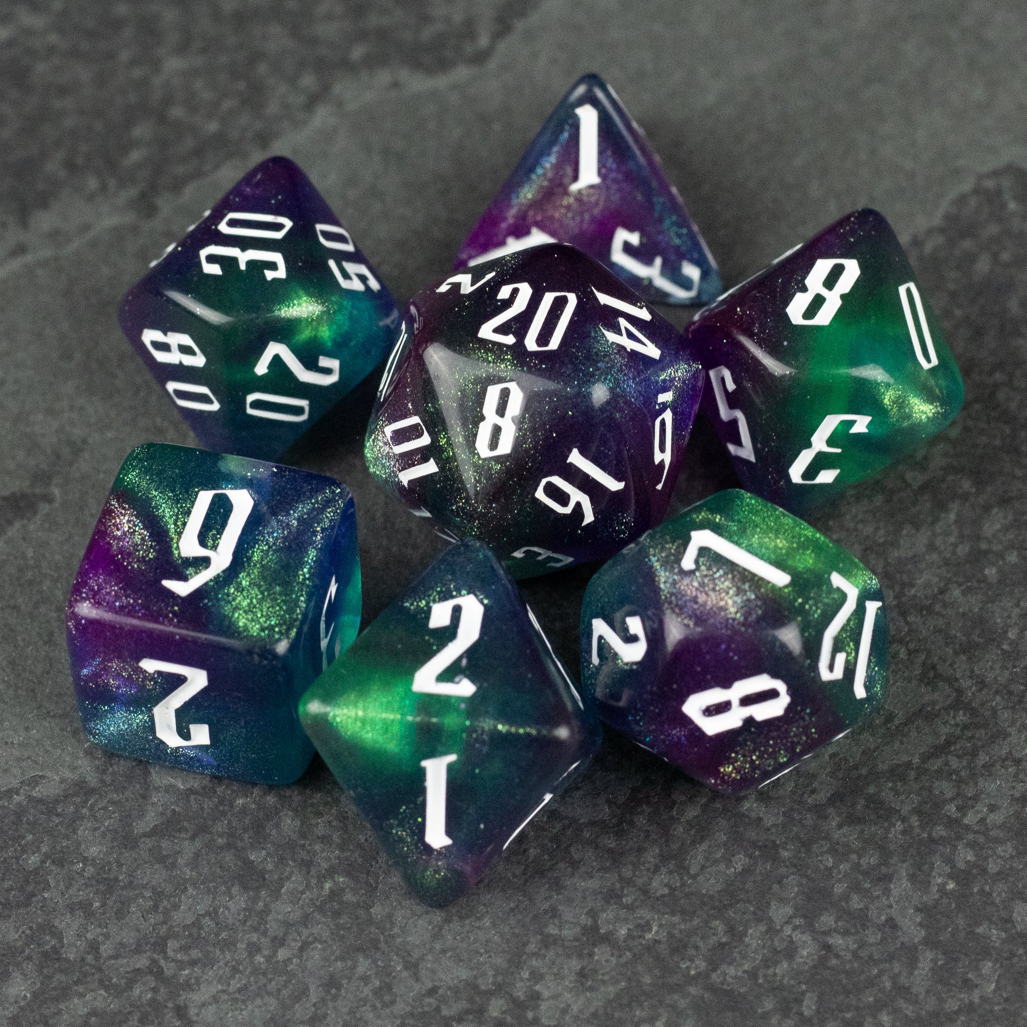 Purple & Green Glitter Dice Set - Seven purple and green glitter polyhedral dice with white numbers on a dark textured surface.