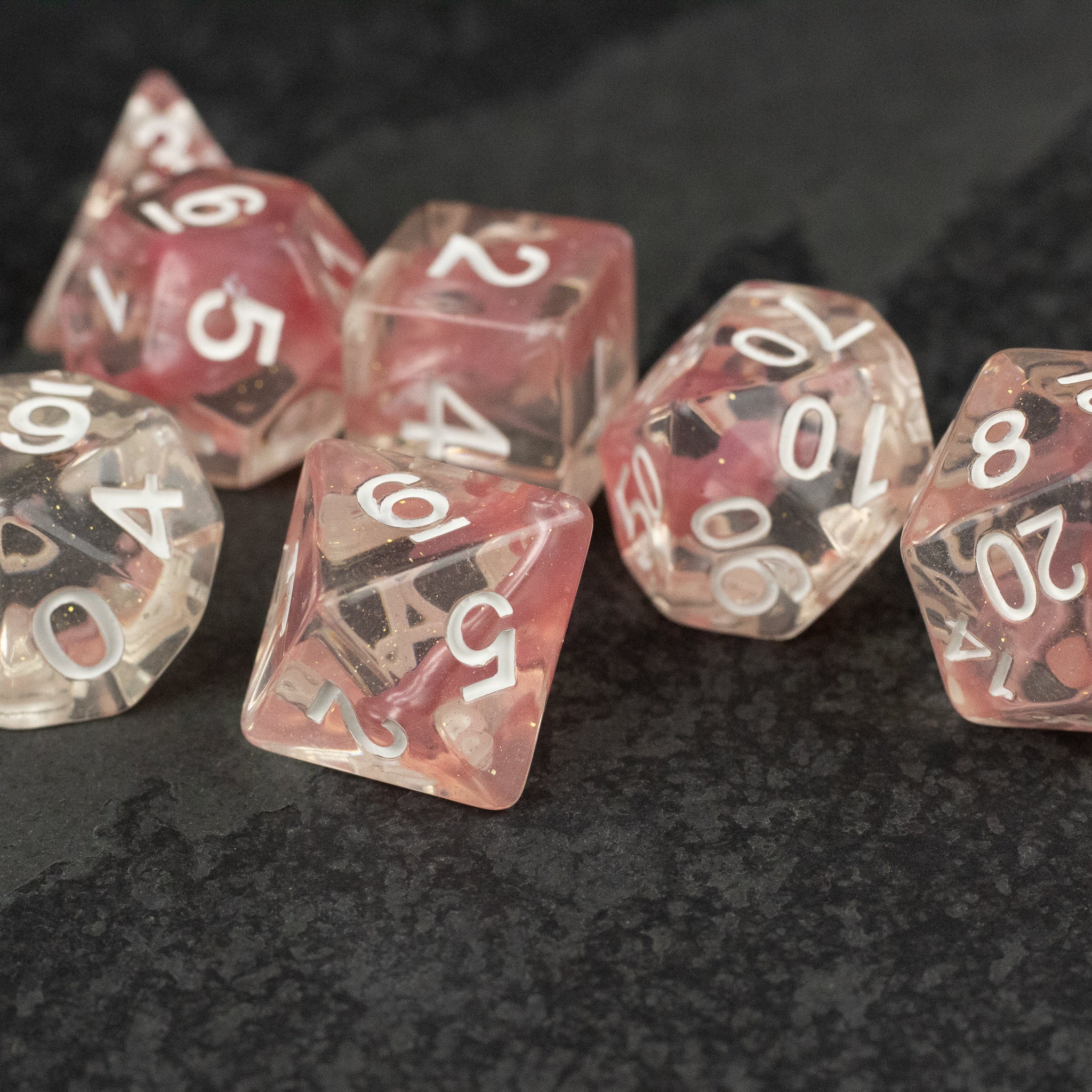Pink Lava Lamp Dice Set - Set of transparent pink polyhedral dice with white numbers, including d20, d12, d10, d8, d6, and d4 on a dark textured.