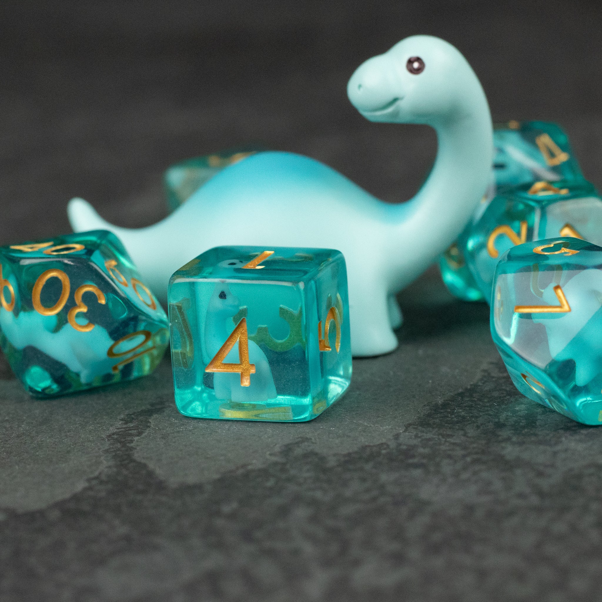 Blue Brontosaurus Dice Set - Translucent blue six-sided dice with gold numbers surrounding a smooth, light blue brontosaurus figurine on a dark surface.