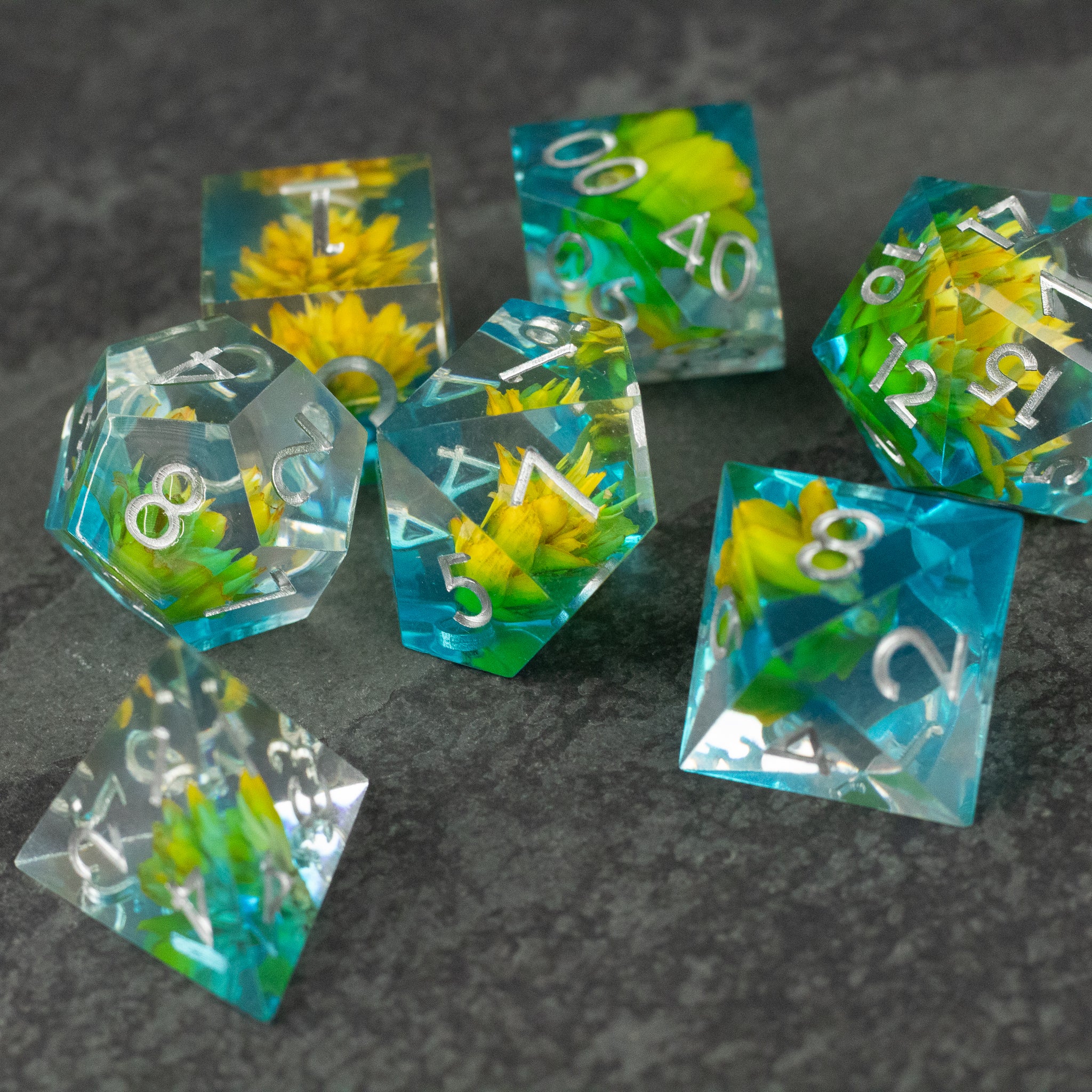 Yellow Flower + Blue Resin Sharp Edge Dice Set - Set of sharp-edged polyhedral dice with translucent blue resin and embedded yellow flowers, white numbering.