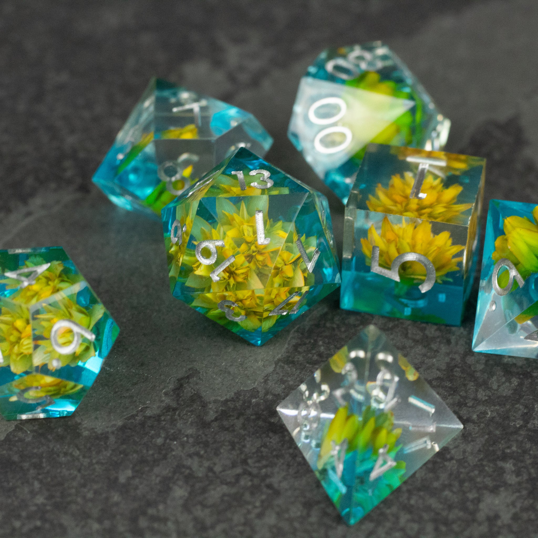 Yellow Flower + Blue Resin Sharp Edge Dice Set - Set of sharp-edged polyhedral dice with translucent blue resin and embedded yellow flowers, white numbering.