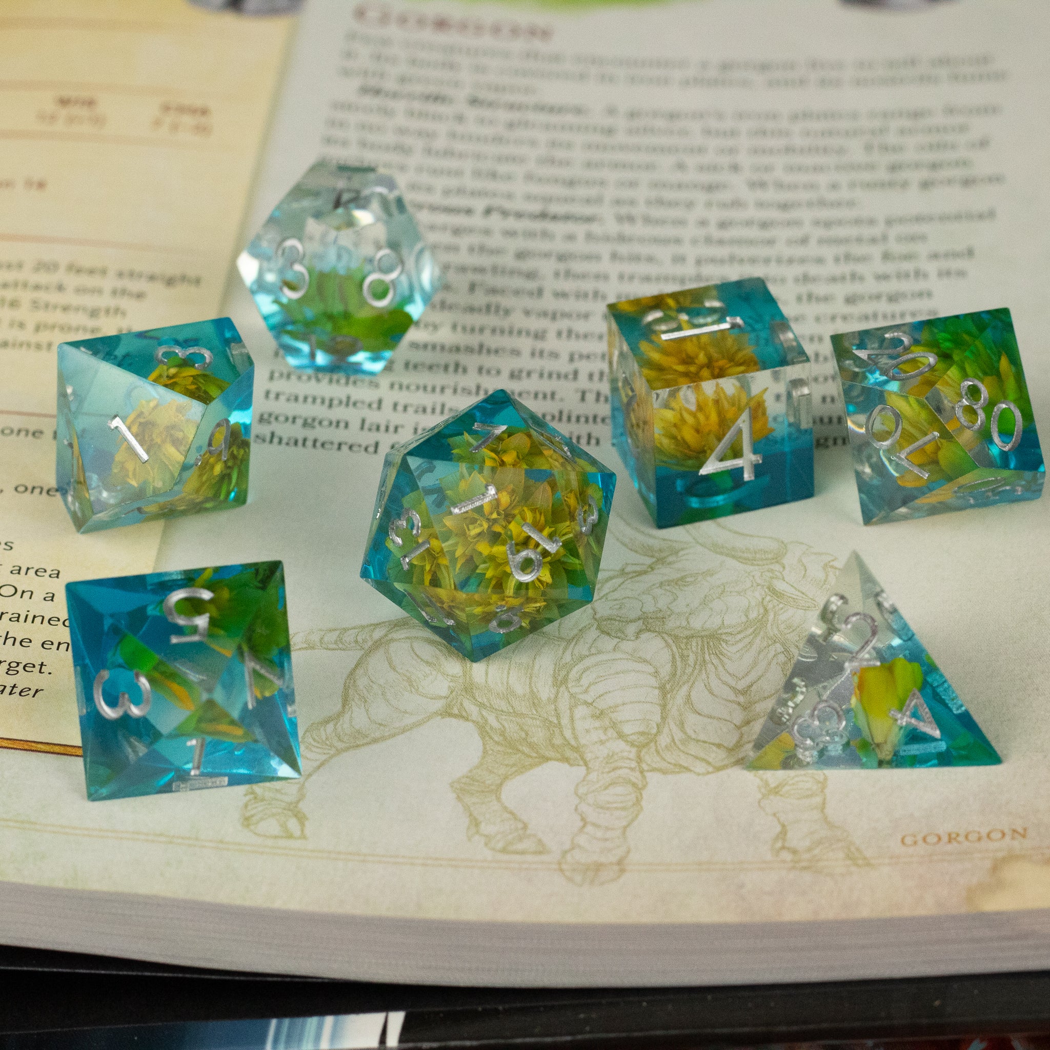 Yellow Flower + Blue Resin Sharp Edge Dice Set - Set of seven sharp edge dice with blue resin and yellow flowers inside on an open book.