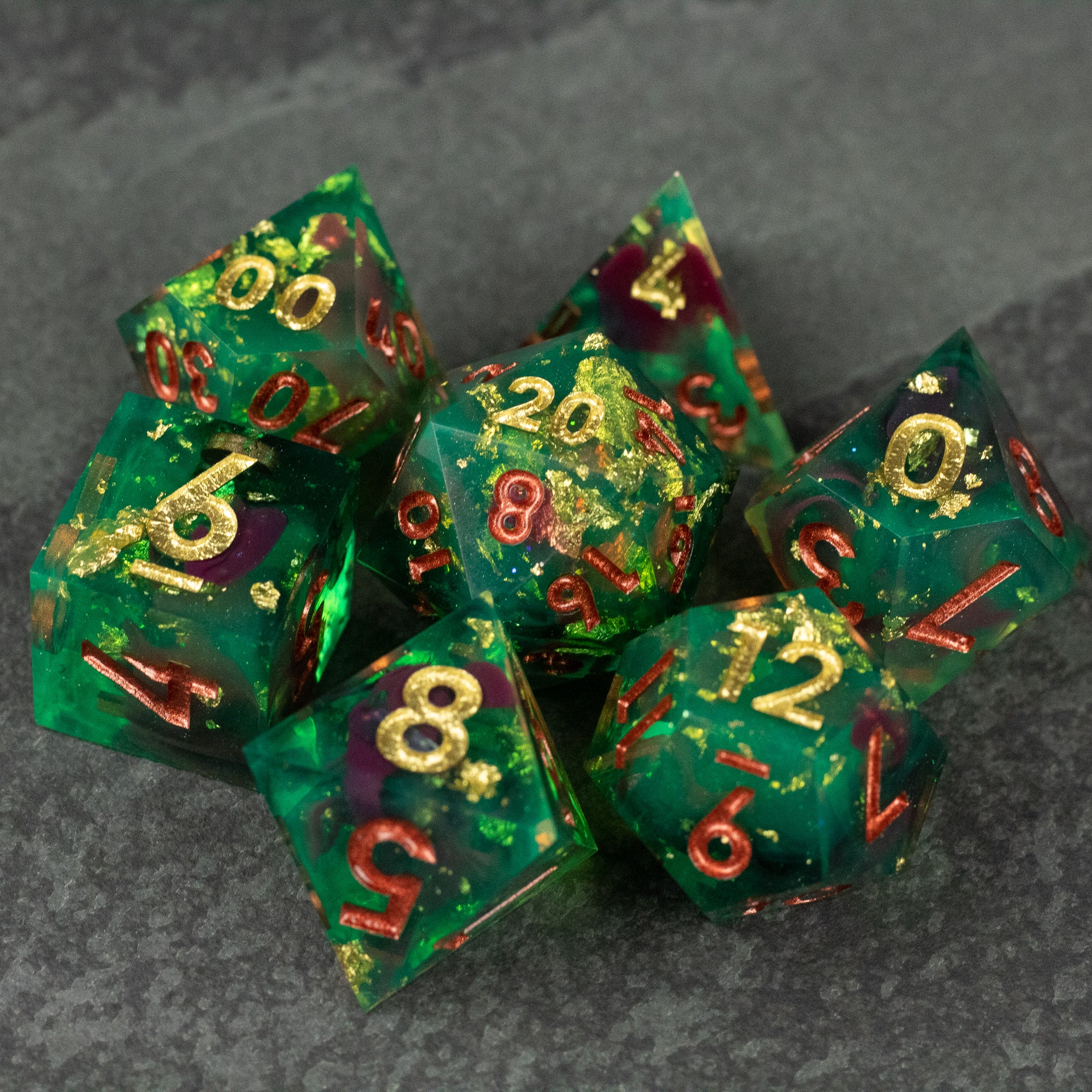 Primal Blood Dice Set - Seven sharp-edged green polyhedral dice with gold and red numbers and embedded gold flakes on dark background.
