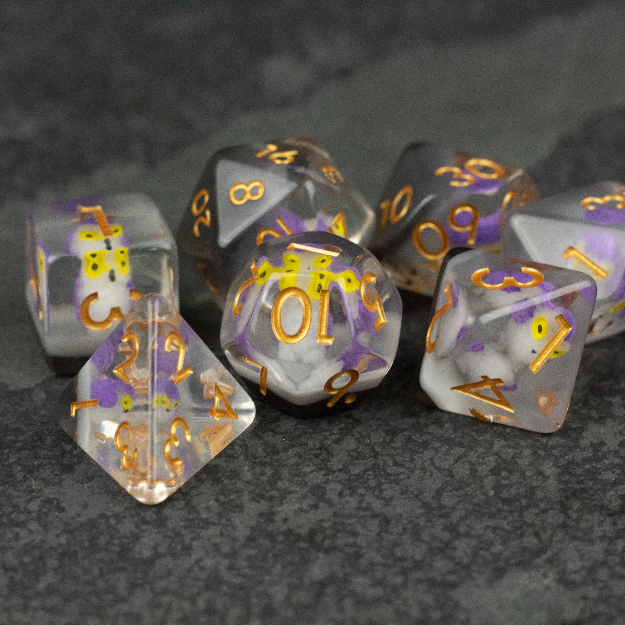 Purple Owl Dice Set - Set of seven translucent purple and clear polyhedral dice with gold numbers on a dark textured surface.