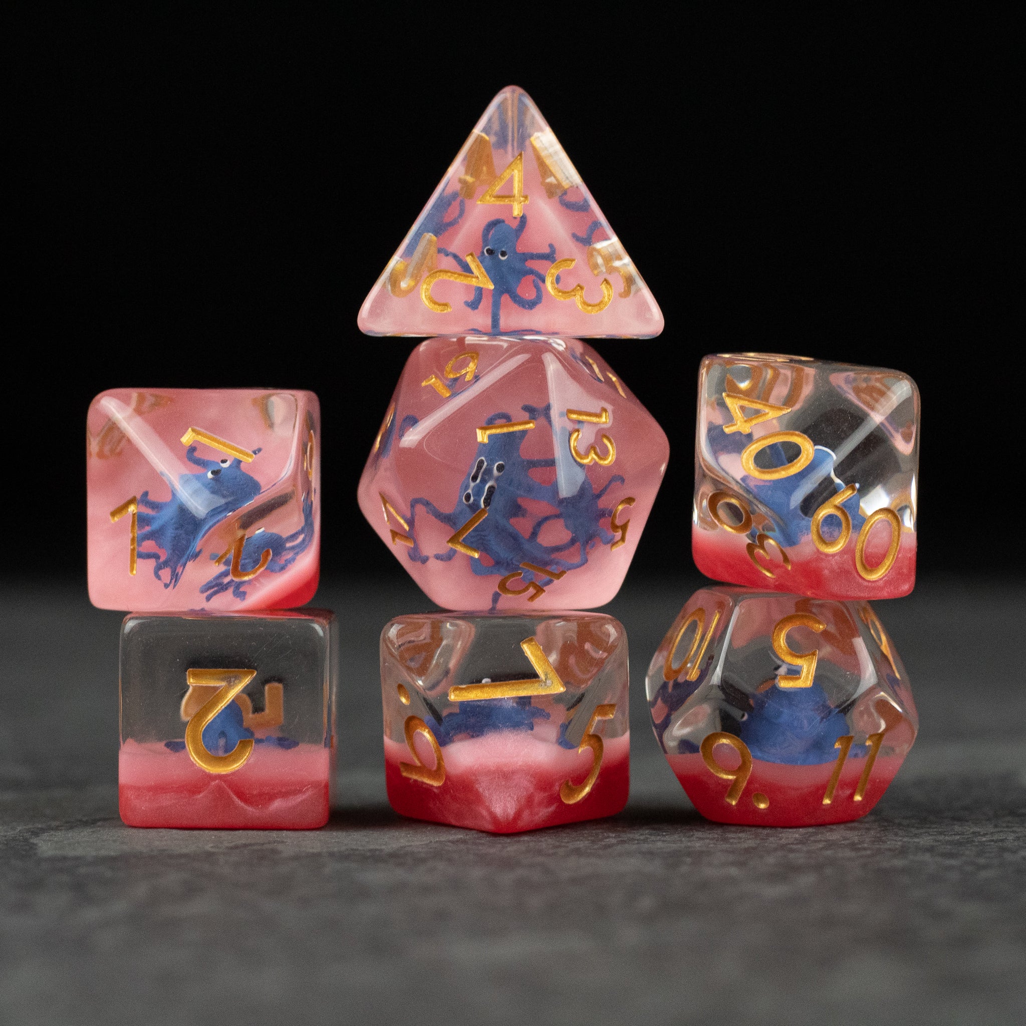 Dark Blue Octopus Dice Set - Set of seven multicolored polyhedral dice with dark blue octopus designs and gold numbers on pink and red backgrounds.