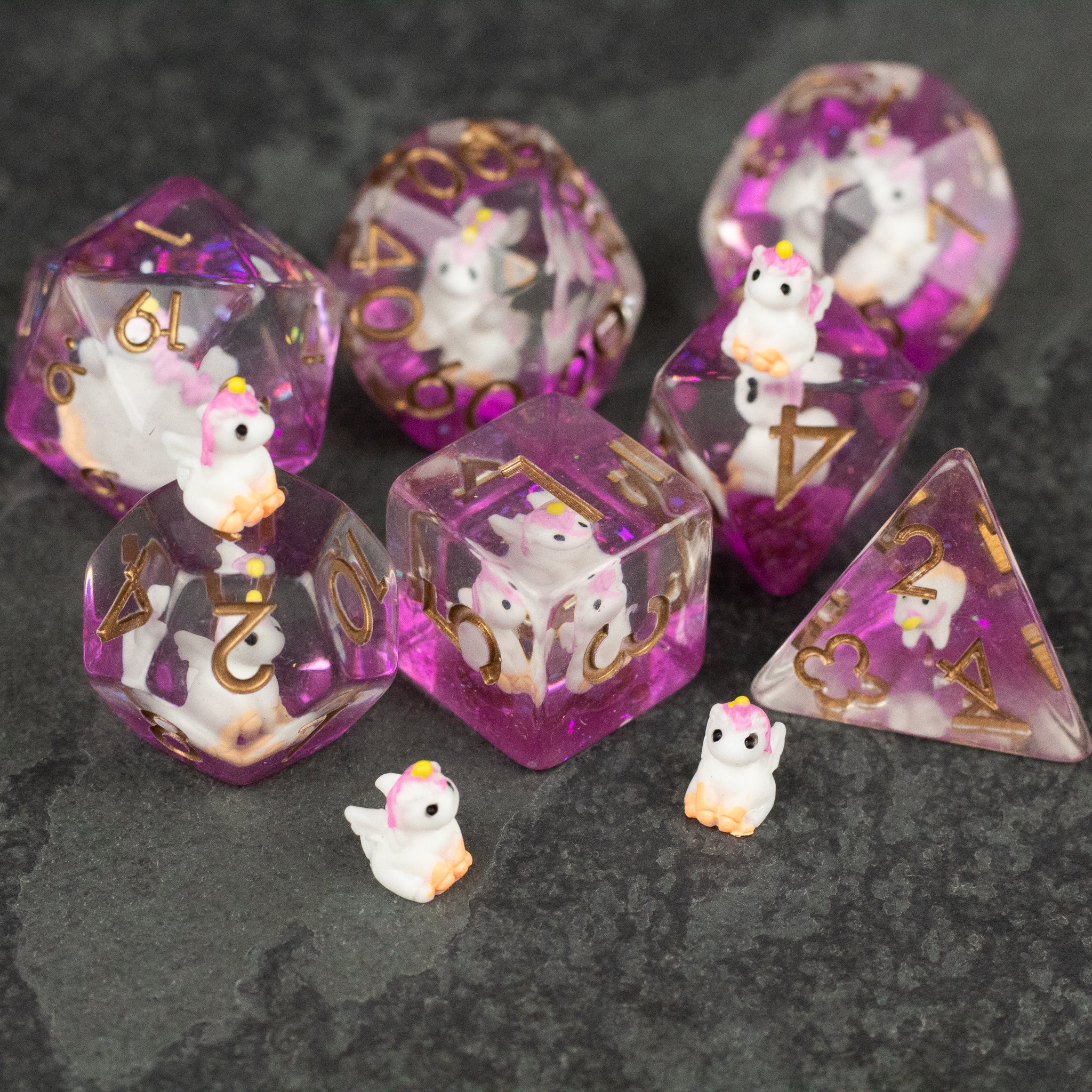 Pink Unicorn Dice Set - Set of seven translucent pink polyhedral dice with gold numbers and small white unicorn figures inside and beside them on a.