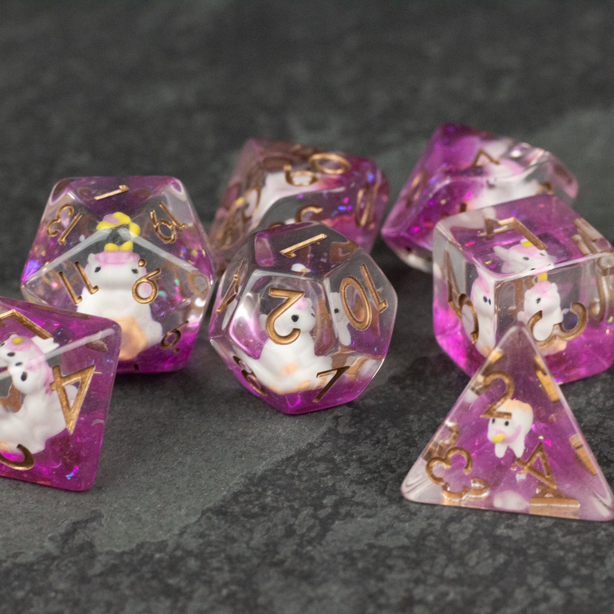 Pink Unicorn Dice Set - Set of translucent pink polyhedral dice with gold numbering and unicorn silhouettes inside on dark textured surface.