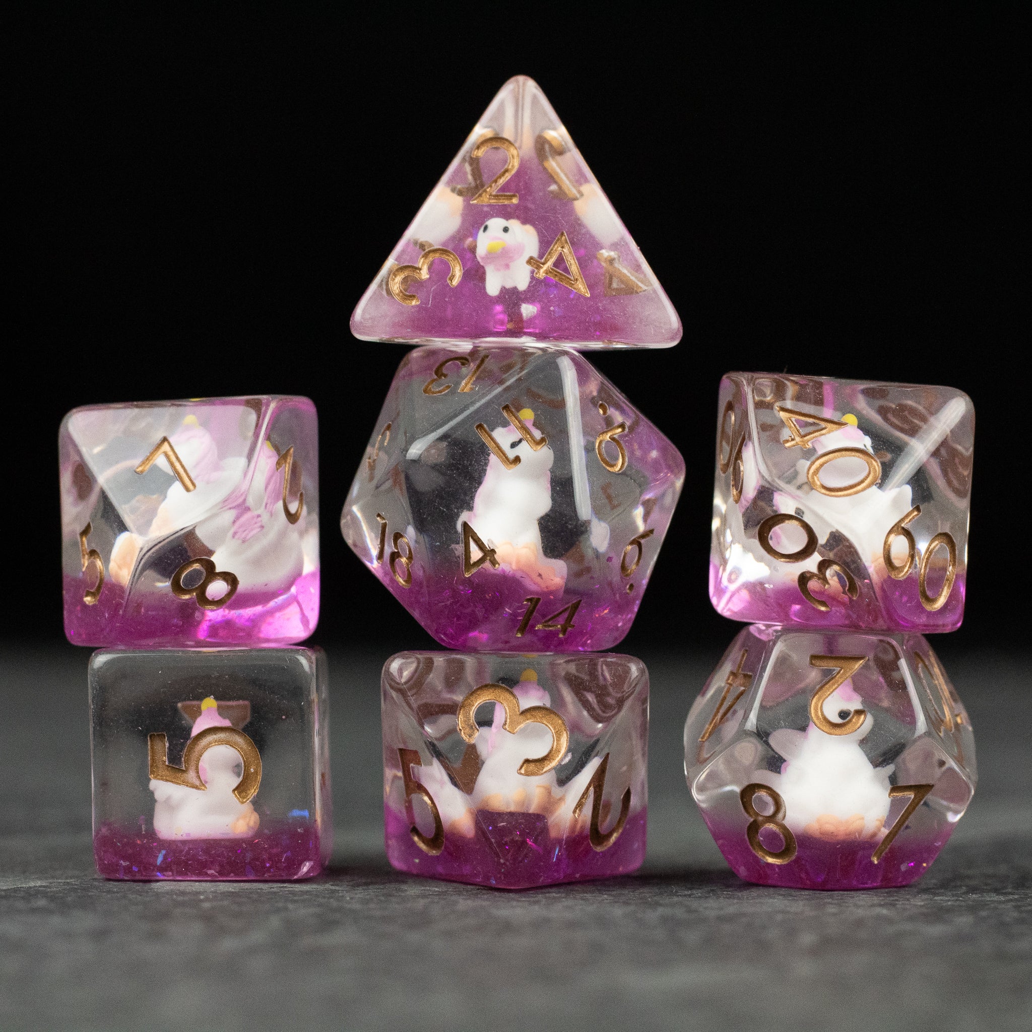 Pink Unicorn Dice Set - Set of seven translucent pink and clear polyhedral dice with gold numbers and unicorn illustrations inside.