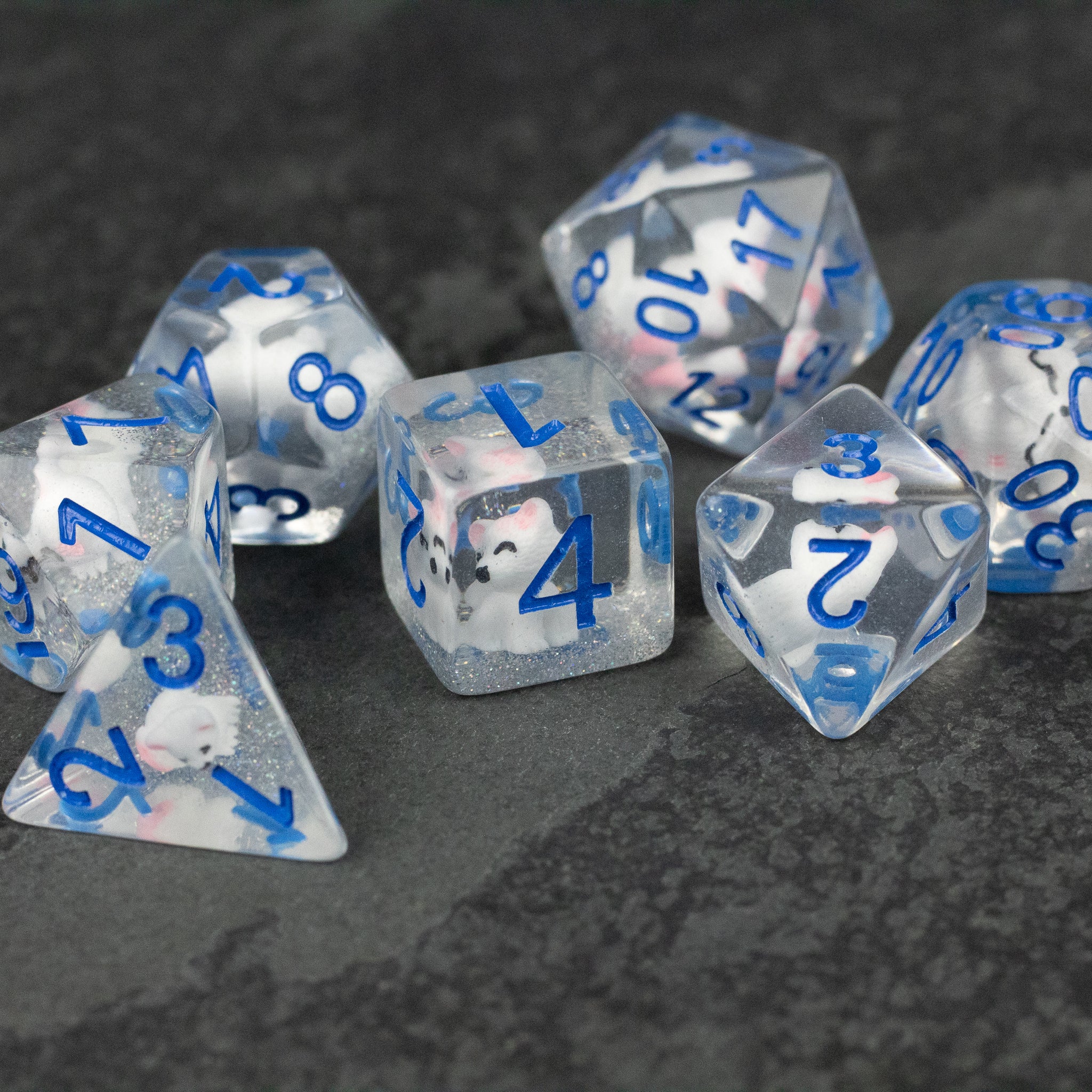Artic Fox Dice Set - Clear polyhedral dice set with blue numbers and small white and black fox designs inside on a dark surface.