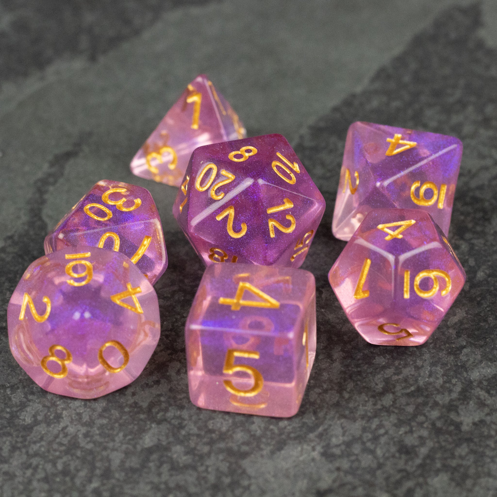 Purple Aura Glitter Dice Set - Seven translucent purple polyhedral dice with gold numbers and a glittery finish on a dark textured surface.