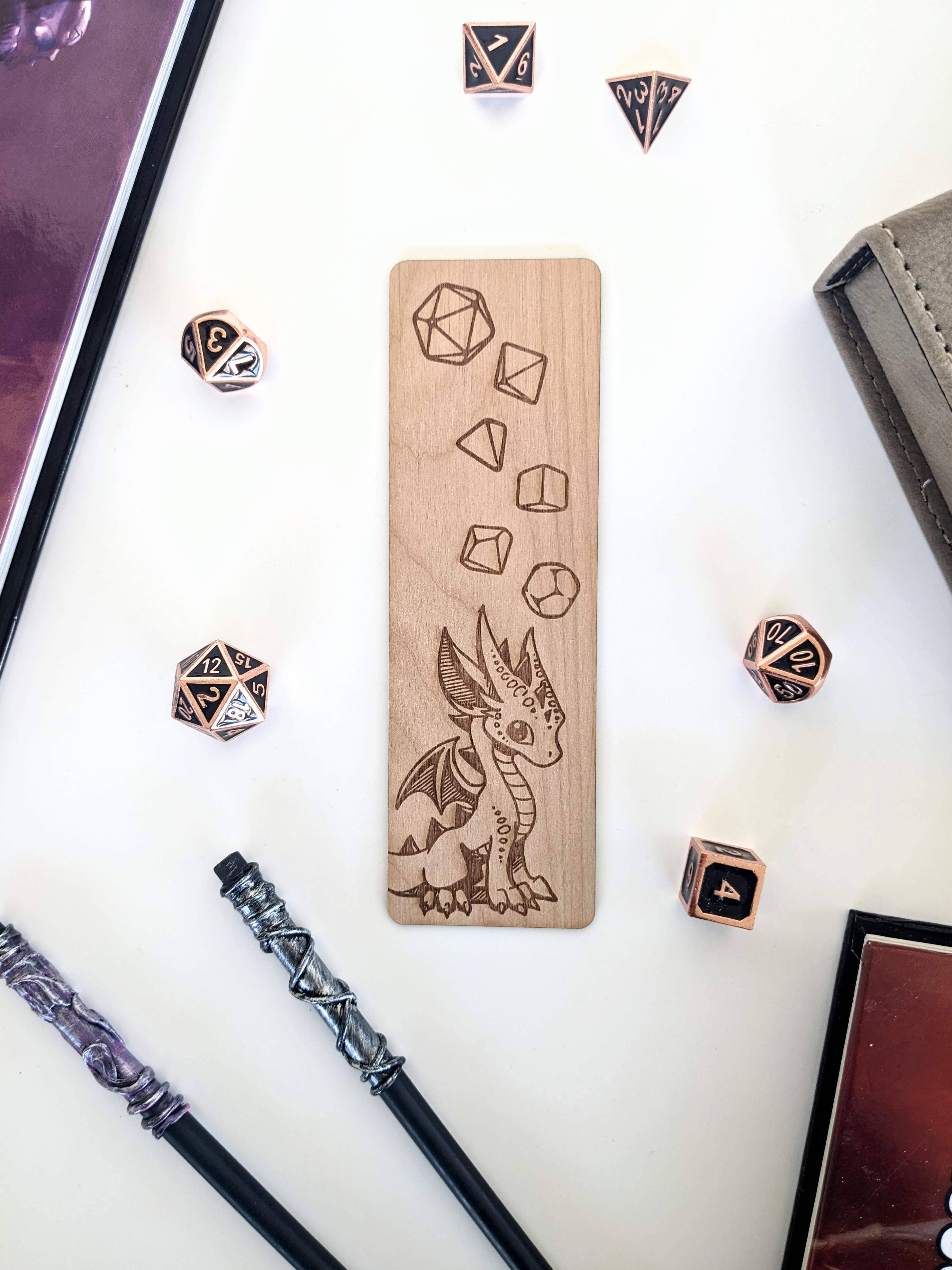 Baby Dice Dragon Wooden Bookmark - Wooden bookmark engraved with a baby dragon and various dice designs, surrounded by black polyhedral dice and two black.