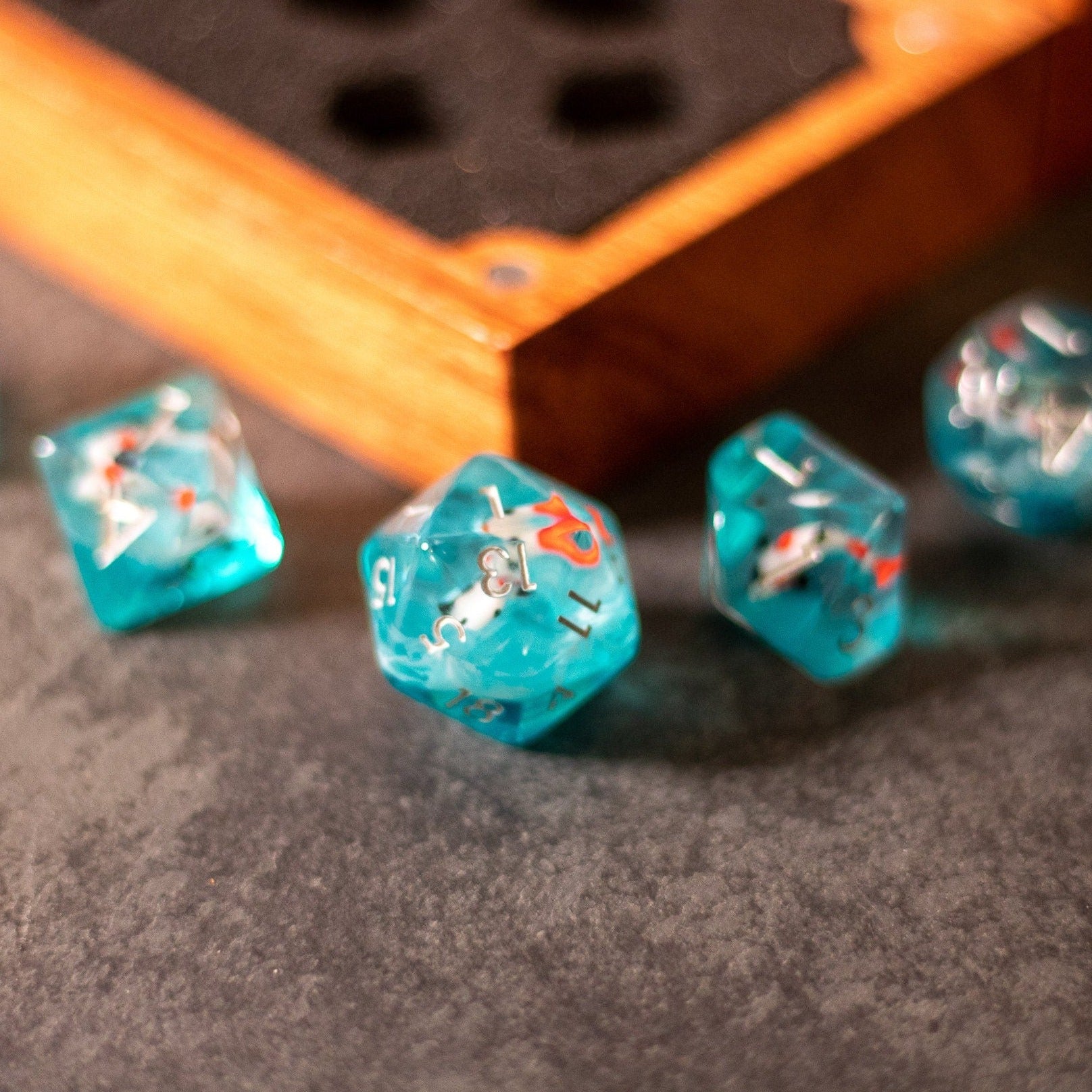 Koi Fish Dice Set - Translucent turquoise polyhedral dice with orange koi fish designs and white numbering, displayed near a wooden dice box.