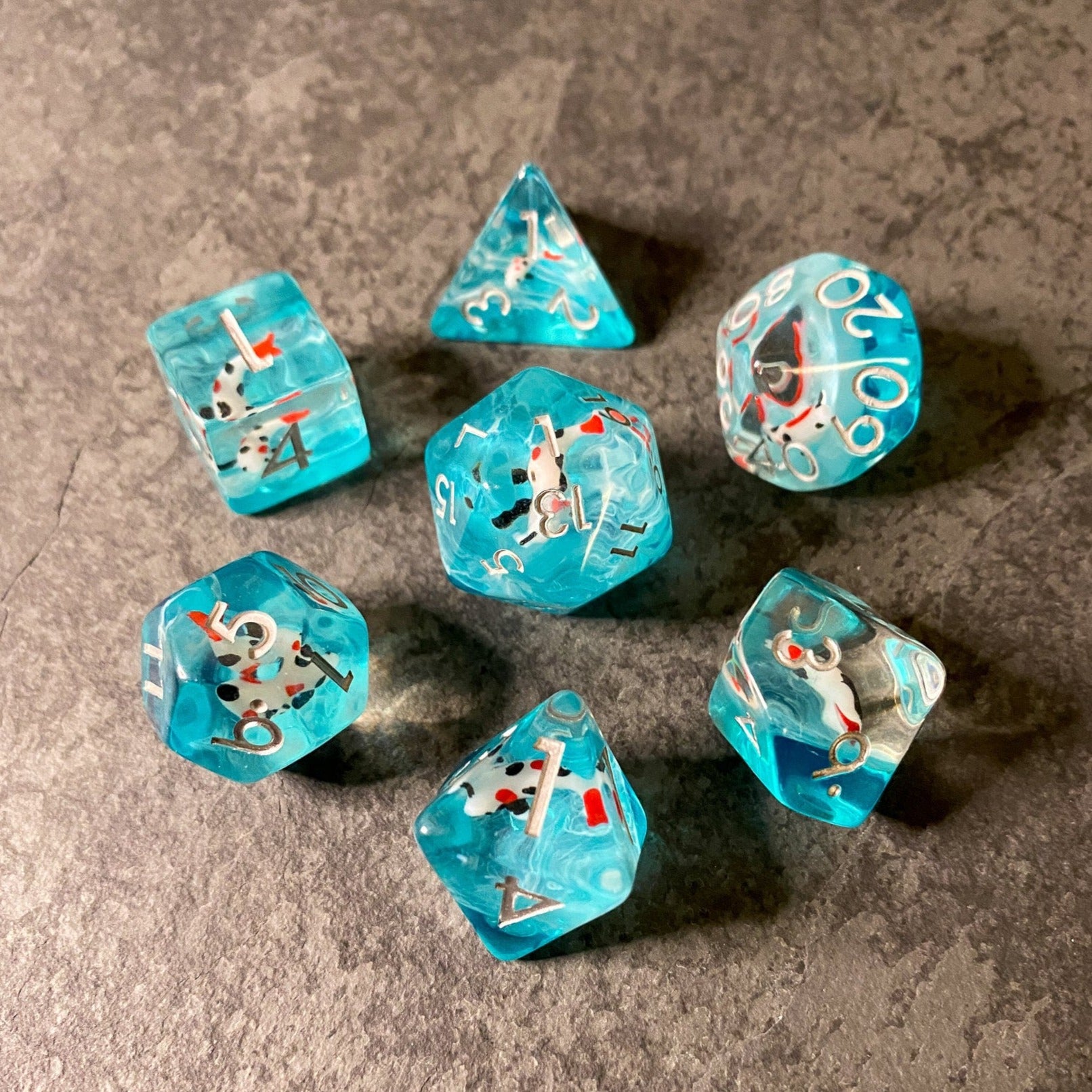 Koi Fish Dice Set - Turquoise polyhedral dice set with koi fish illustrations and white numbering on gray surface.
