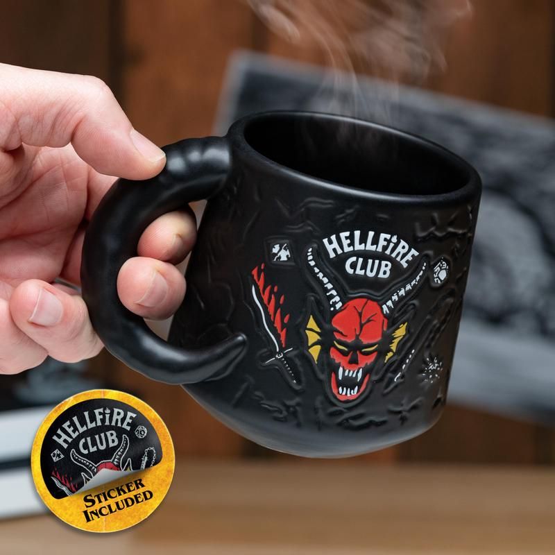 Hellfire Club Mug - Black ceramic mug with Hellfire Club logo featuring a red horned demon and skulls, held in hand with steam rising.