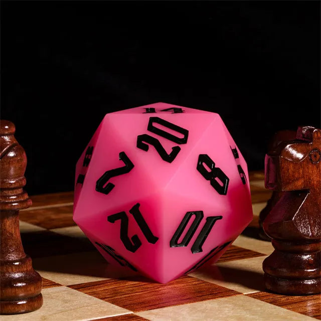 Giant Bouncy D20 - Glow In The Dark Pink - Large pink twenty-sided die with bold black numbers, glowing material, resting on a wooden chessboard.