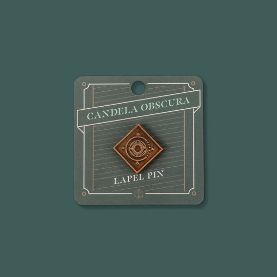 Candela Obscura Lapel Pin - Copper-toned diamond-shaped lapel pin with concentric circle design on dark green backing card.