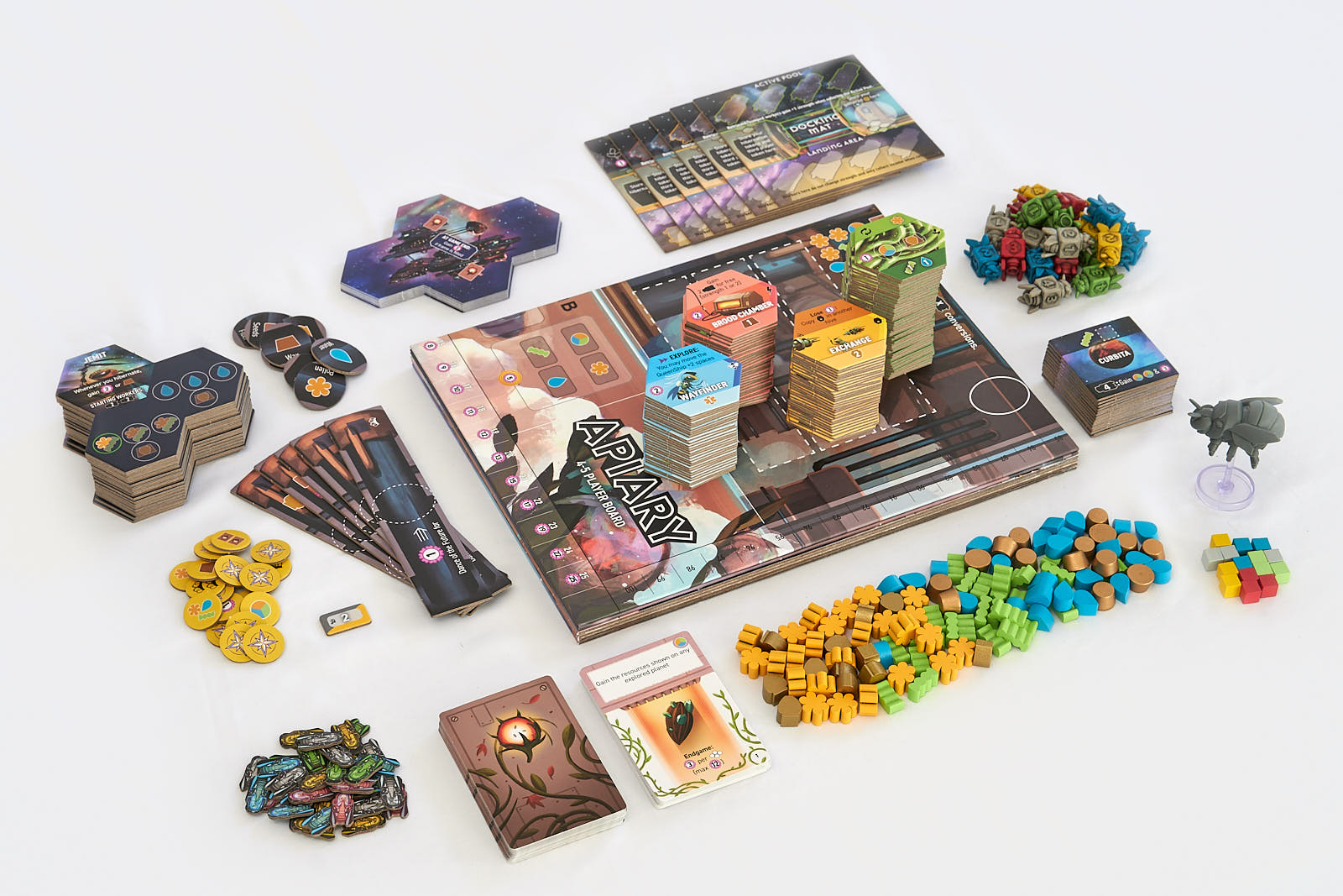 Apiary - Board game components with colorful hexagonal tiles, wooden bee and flower tokens, cards, and various game pieces arranged.