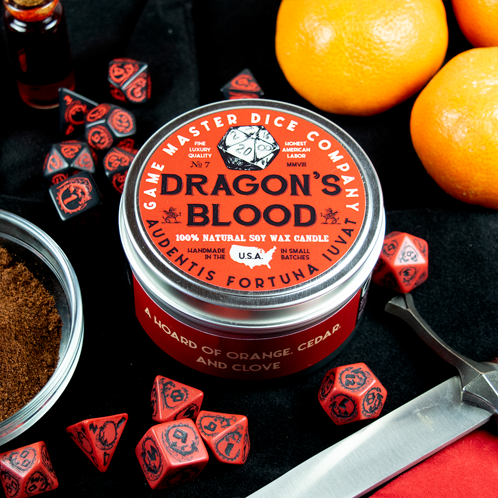 Dragon's Blood Gaming Candle - Red glass jar candle with orange and black label reading "Dragon's Blood," surrounded by red and black gaming dice and an.