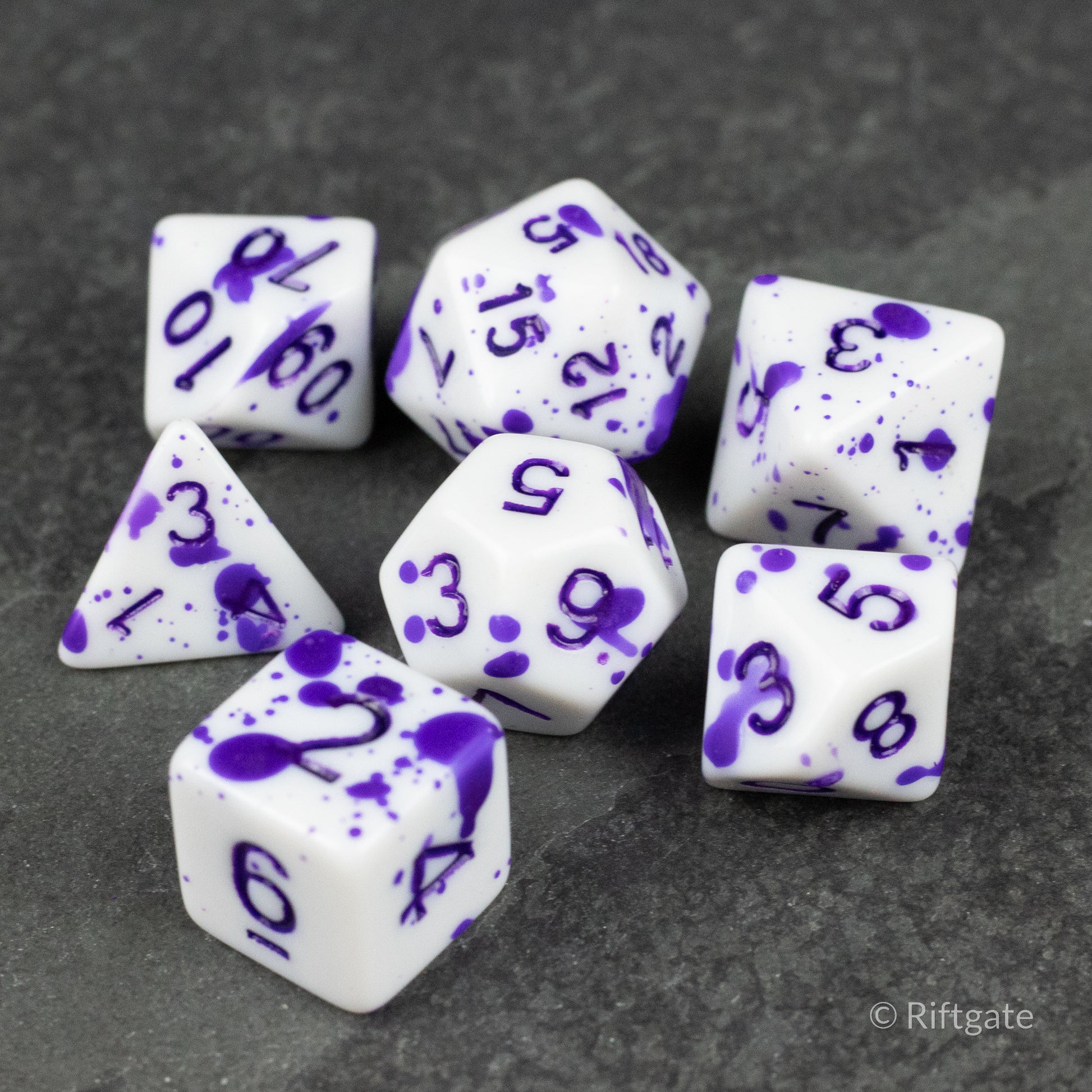 Color Change Purple Splatter Dice Set - Seven white polyhedral dice with purple splatter paint and purple numbers, including d4, d6, d8, d10, d12, and d20 shapes.